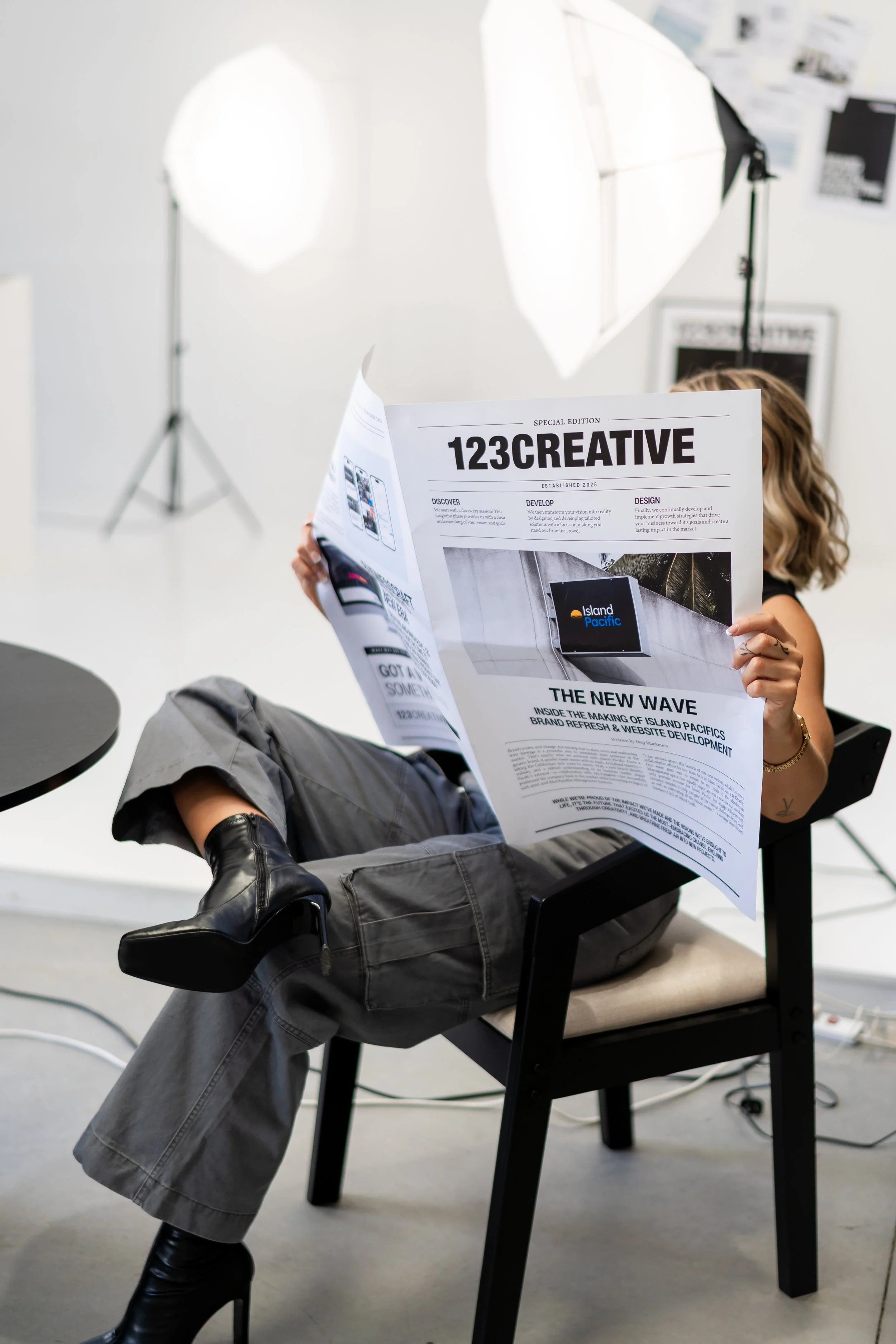 Person sitting on a chair reading a newspaper in a photography studio with professional lighting equipment in the background.