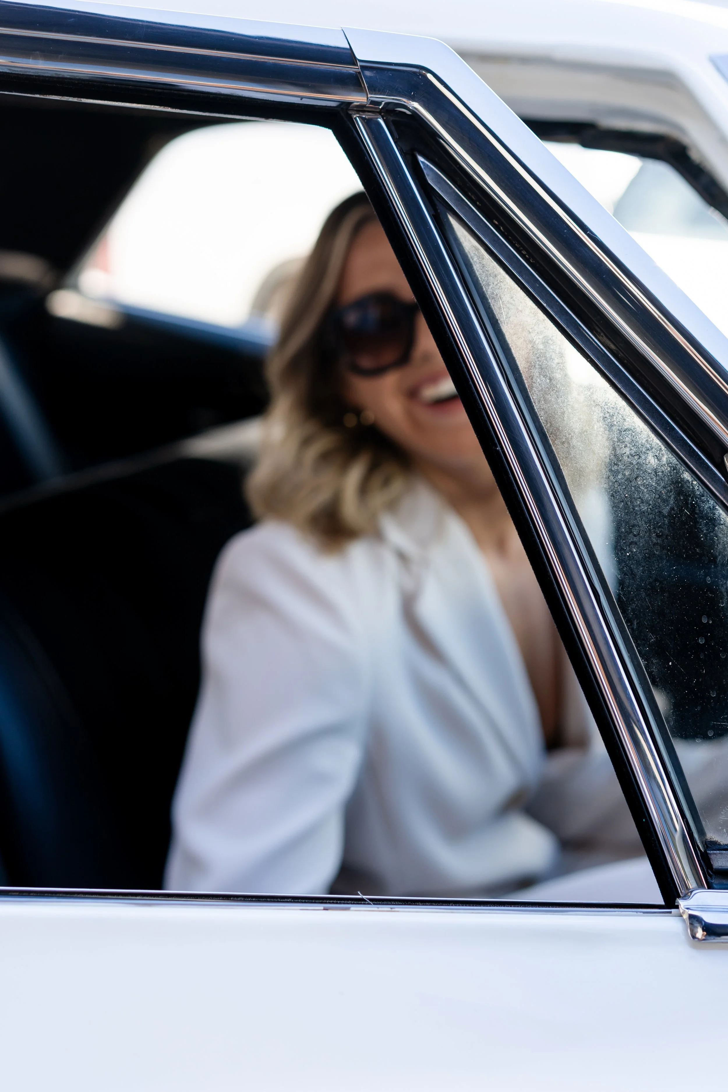 A woman with blonde, wavy hair, wearing large sunglasses and a white blazer, sitting inside a white car, smiling.