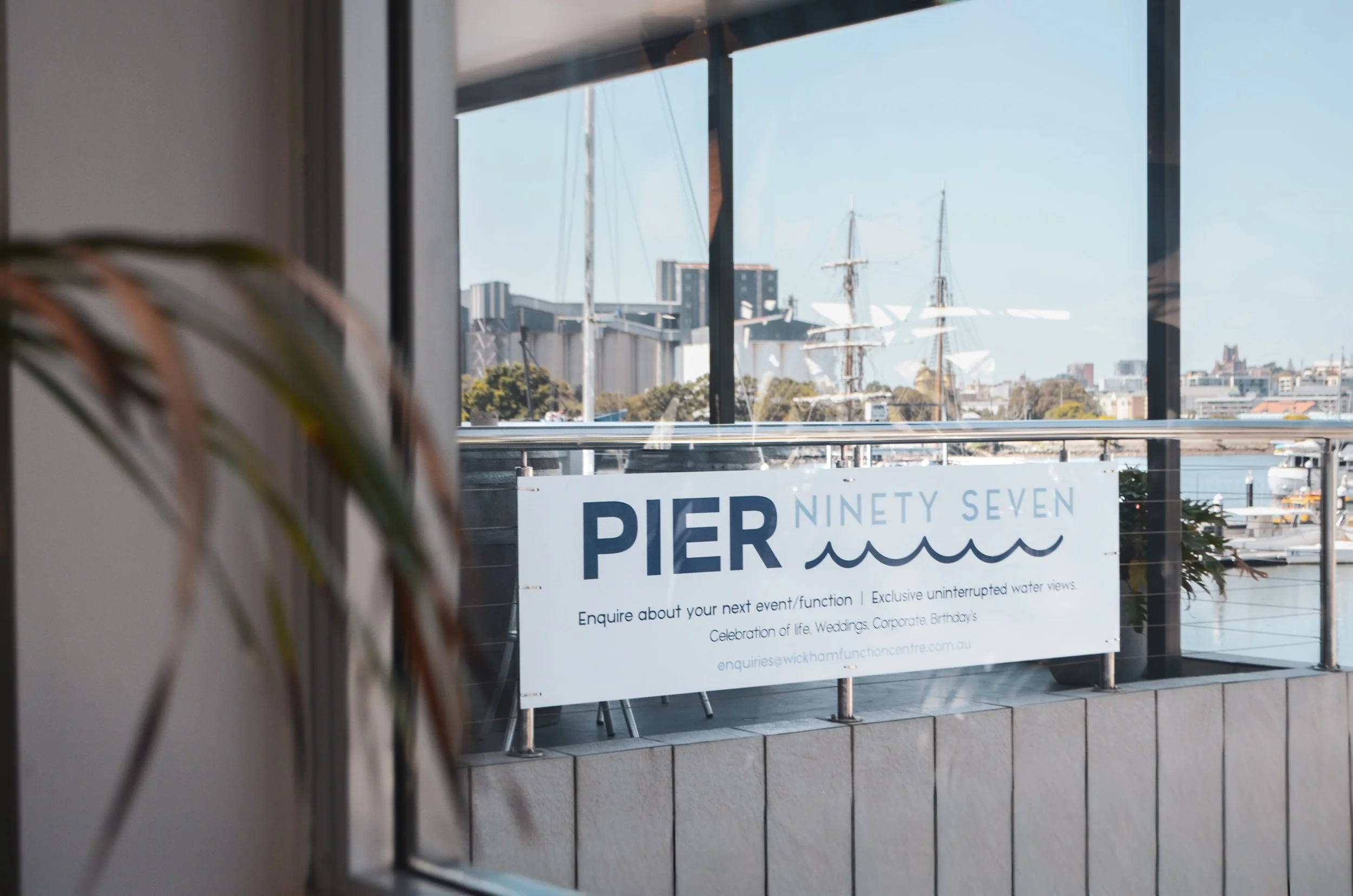 View of a harbor seen through large windows, with sailboats and yachts docked, and a sign that reads 'Pier Ninety Seven' inviting inquiries about events or functions with water views.