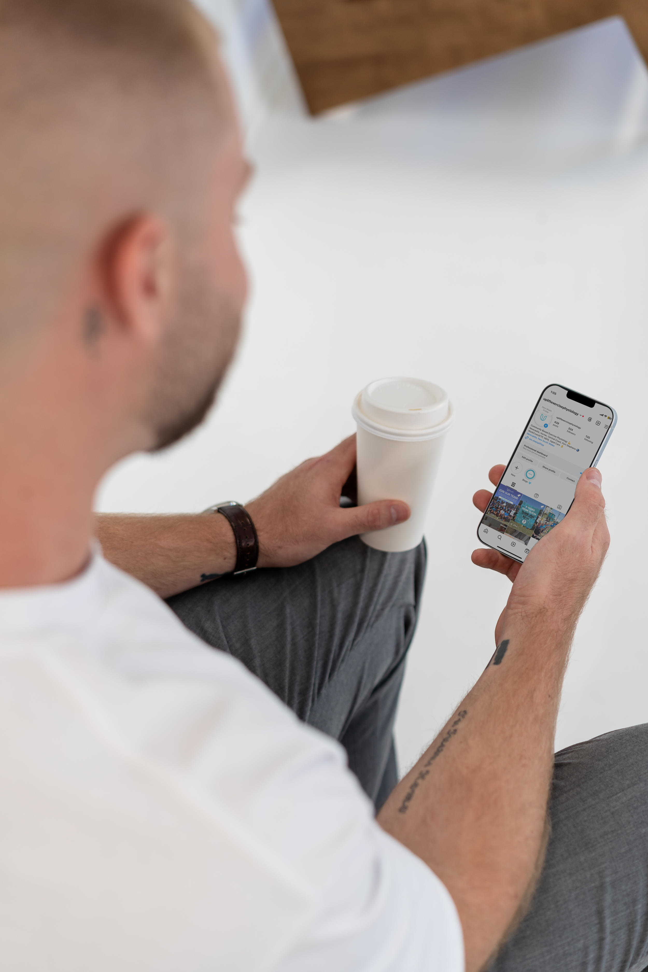 A man with tattooed arms and a wristwatch sitting on a white surface, holding a white coffee cup in one hand and a smartphone in the other. He is looking at the phone, which displays social media content.