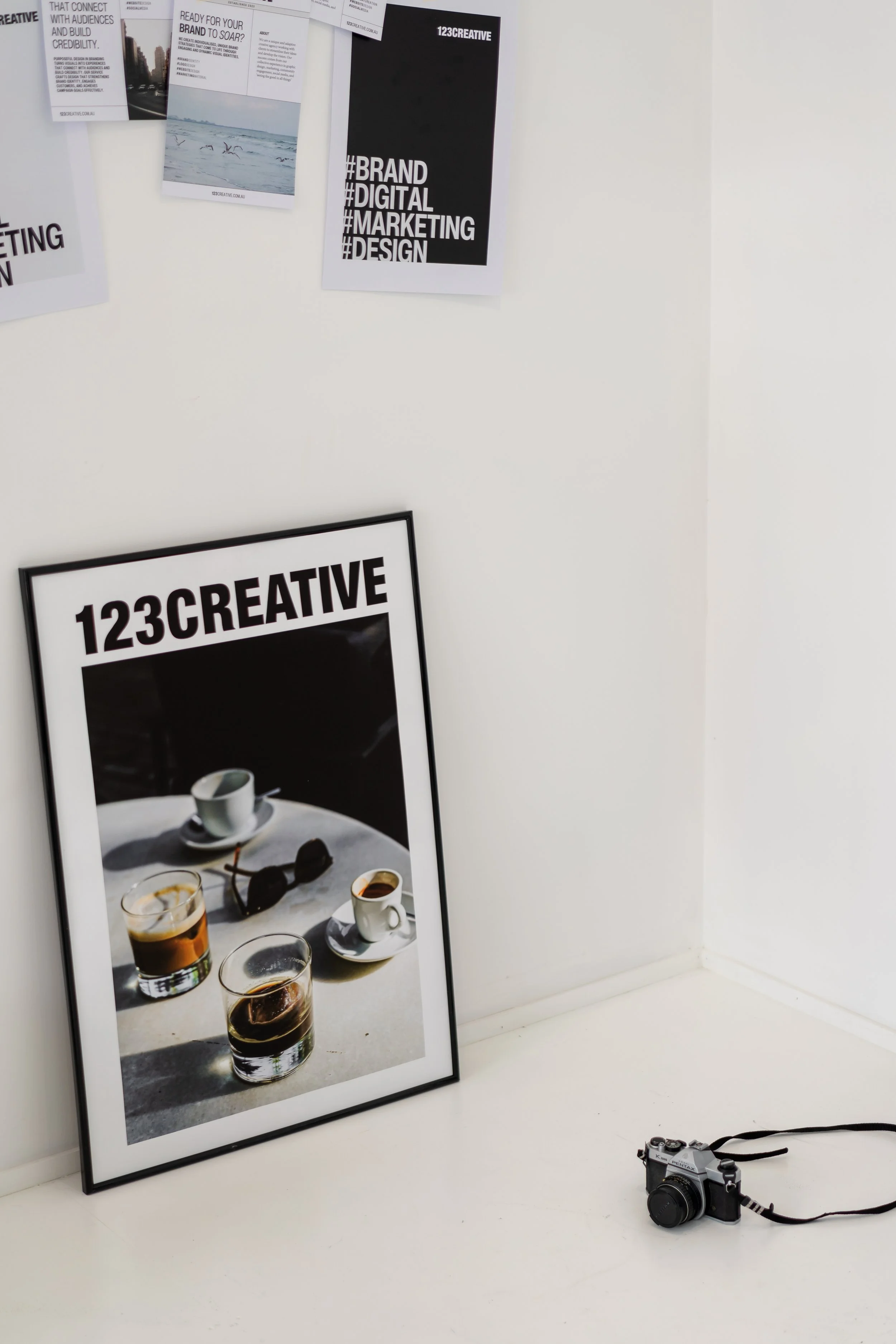 A large framed poster with the text '123CREATIVE' leans against a white wall, featuring a photograph of two cups of coffee, a pair of sunglasses, and two glasses of dark beverages. Several smaller printed sheets about branding, marketing, and design are taped or pinned to the wall above the poster. A digital camera with a strap is placed on the white floor next to the poster.