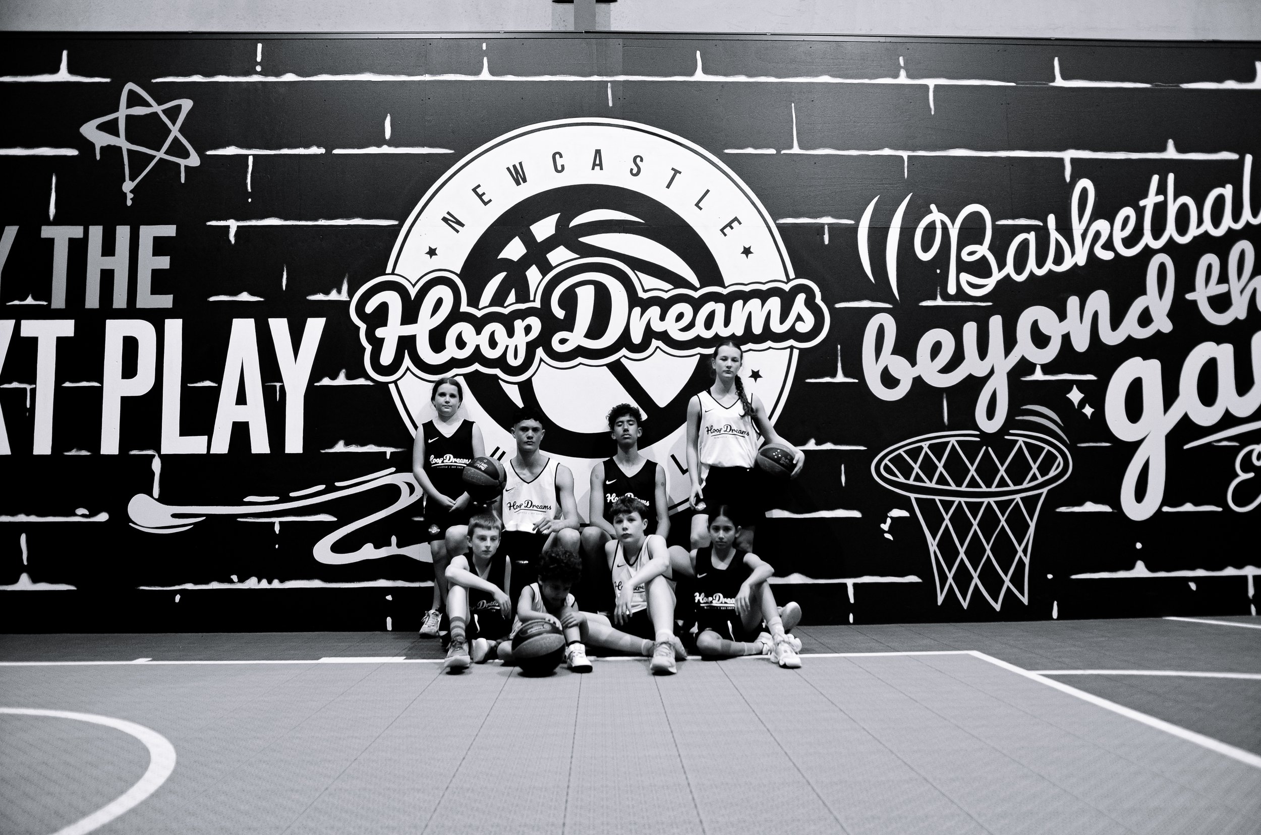 Group of young basketball players posing for a photo in front of a large mural with basketball-themed artwork and slogans, on an indoor court.