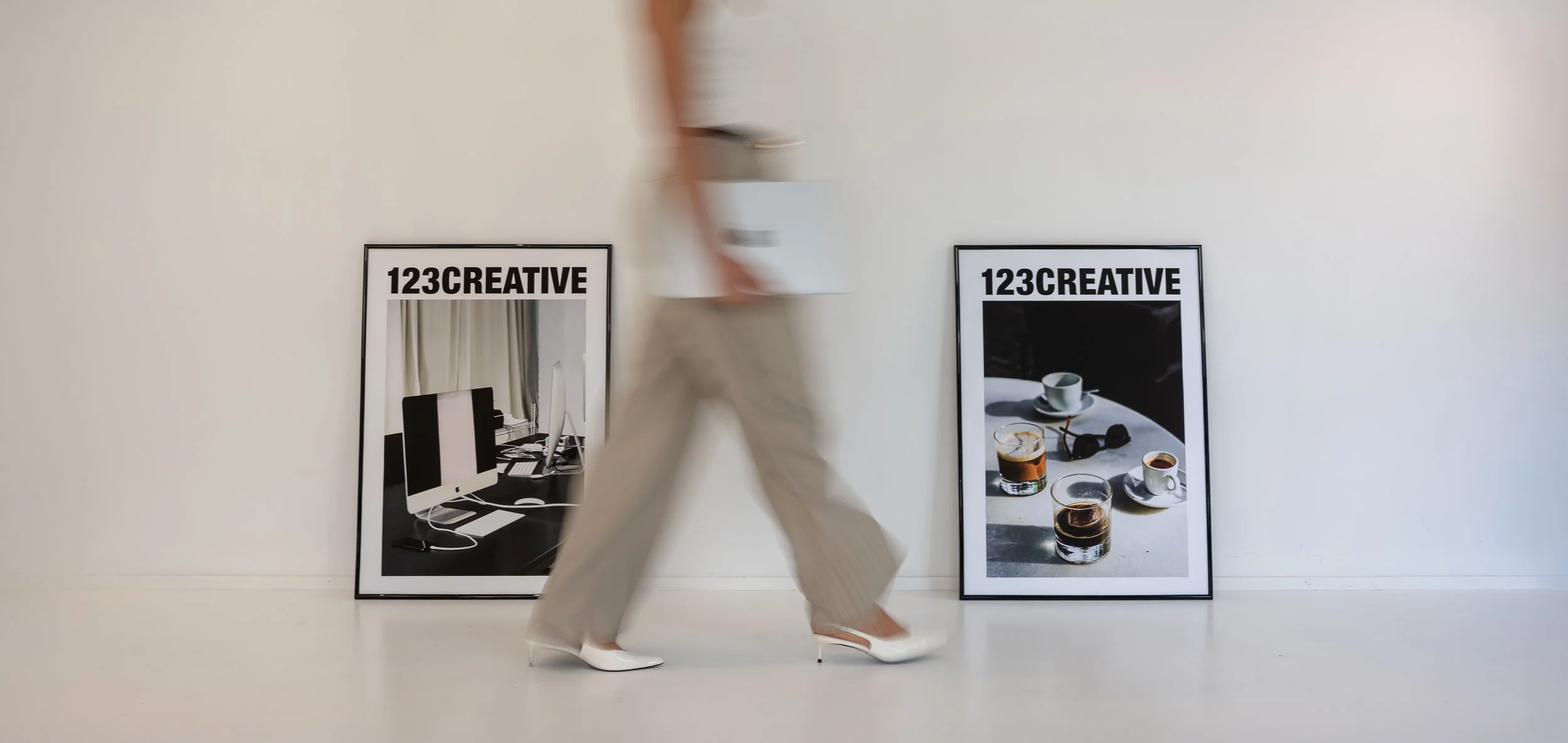 A person walking past two framed black and white posters that say '123CREATIVE', one showing a workspace with an iMac computer, the other showing a table with coffee cups and glasses of water.