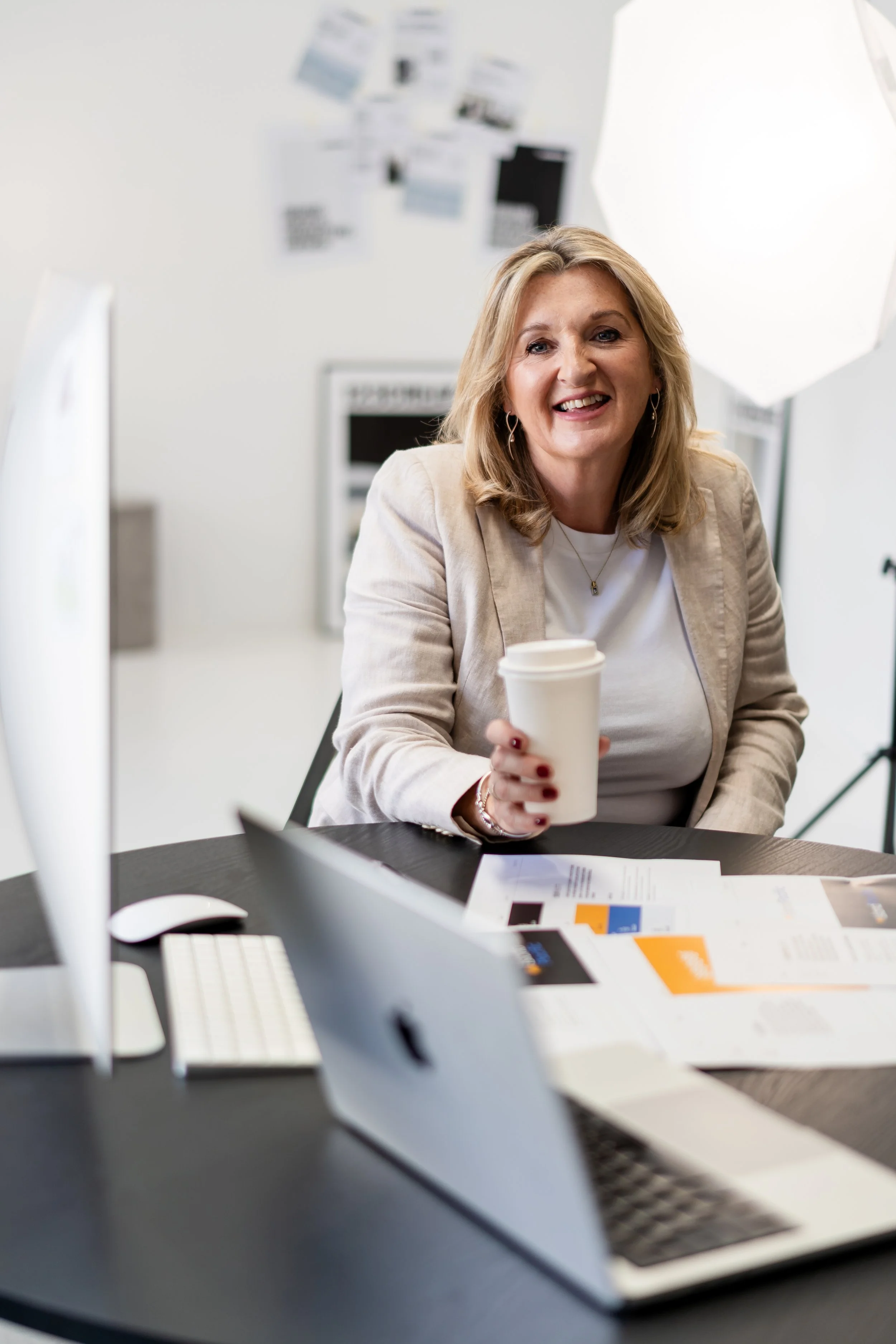 A woman with shoulder-length blonde hair smiling at the camera, sitting at a desk in a bright office. She is holding a white coffee cup, and there are papers, a desktop computer, and a laptop on the desk. The background shows a white wall with pictures and office equipment.