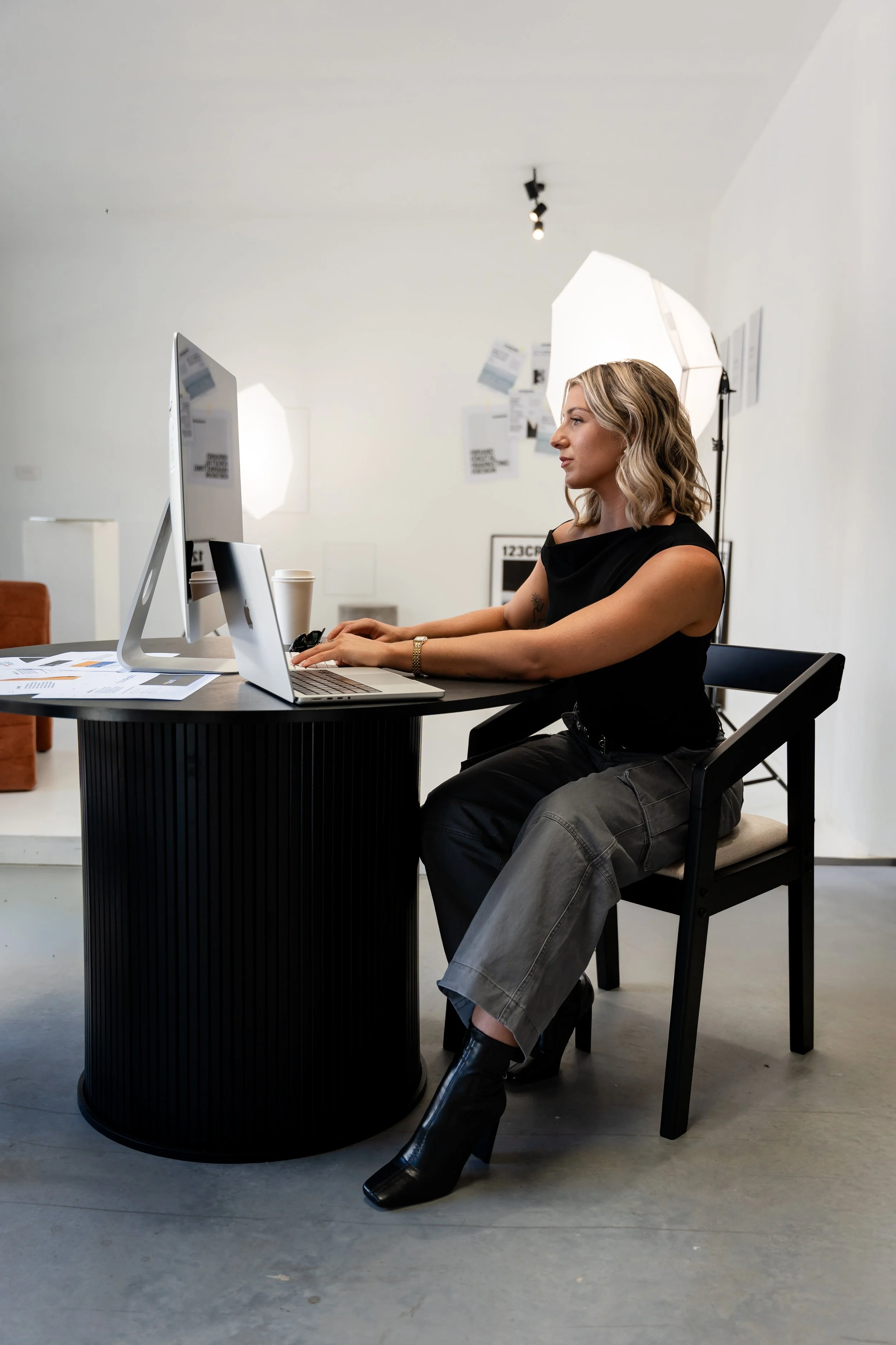 A woman working at a desk in a photography studio with white walls, professional lighting, and a computer.