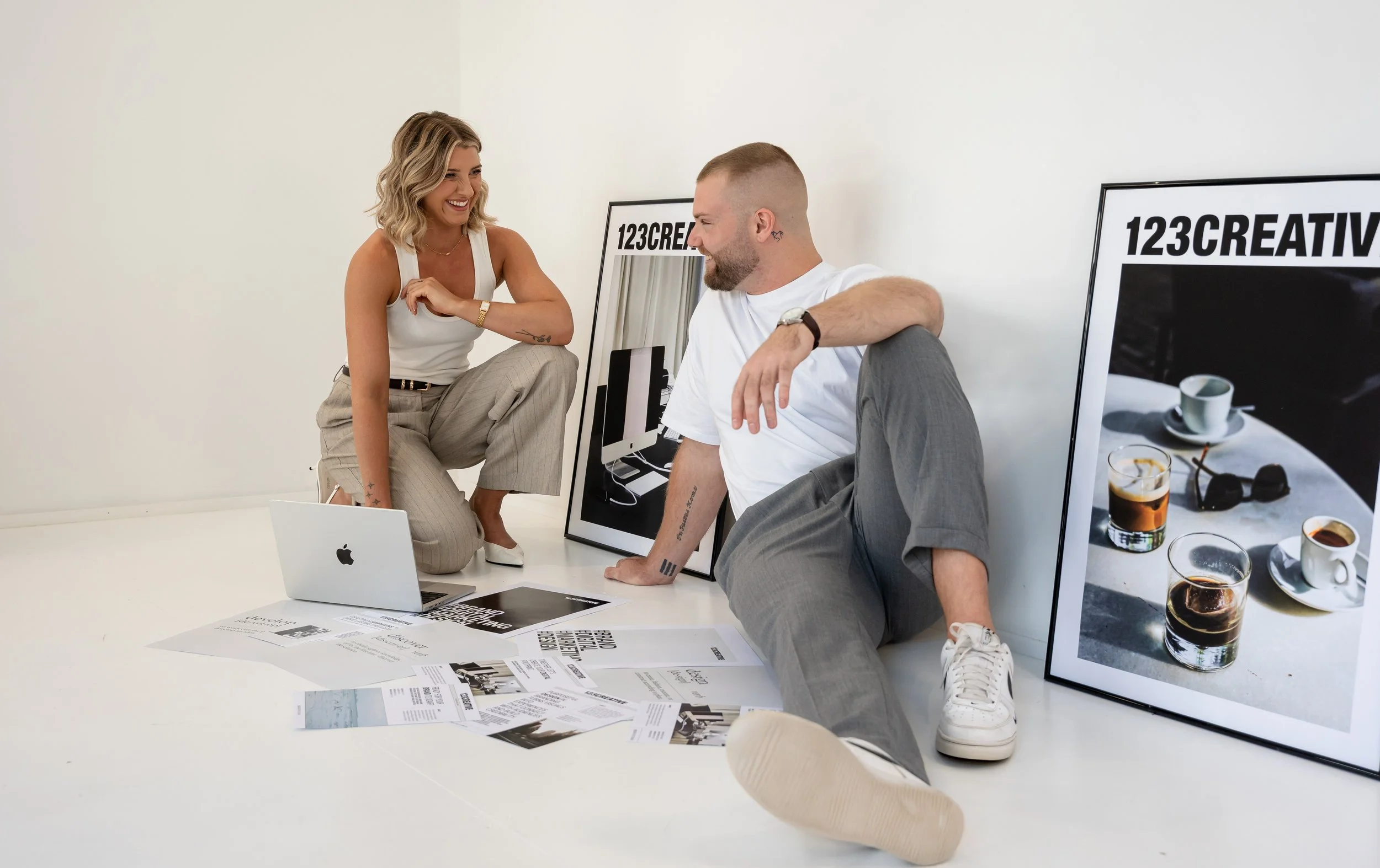 A woman and a man are sitting on the floor, smiling and talking. The woman is wearing a white sleeveless top and beige pants, while the man is dressed in a white t-shirt and gray pants. There are posters, printed materials, a laptop, coffee cups, and sunglasses around them.