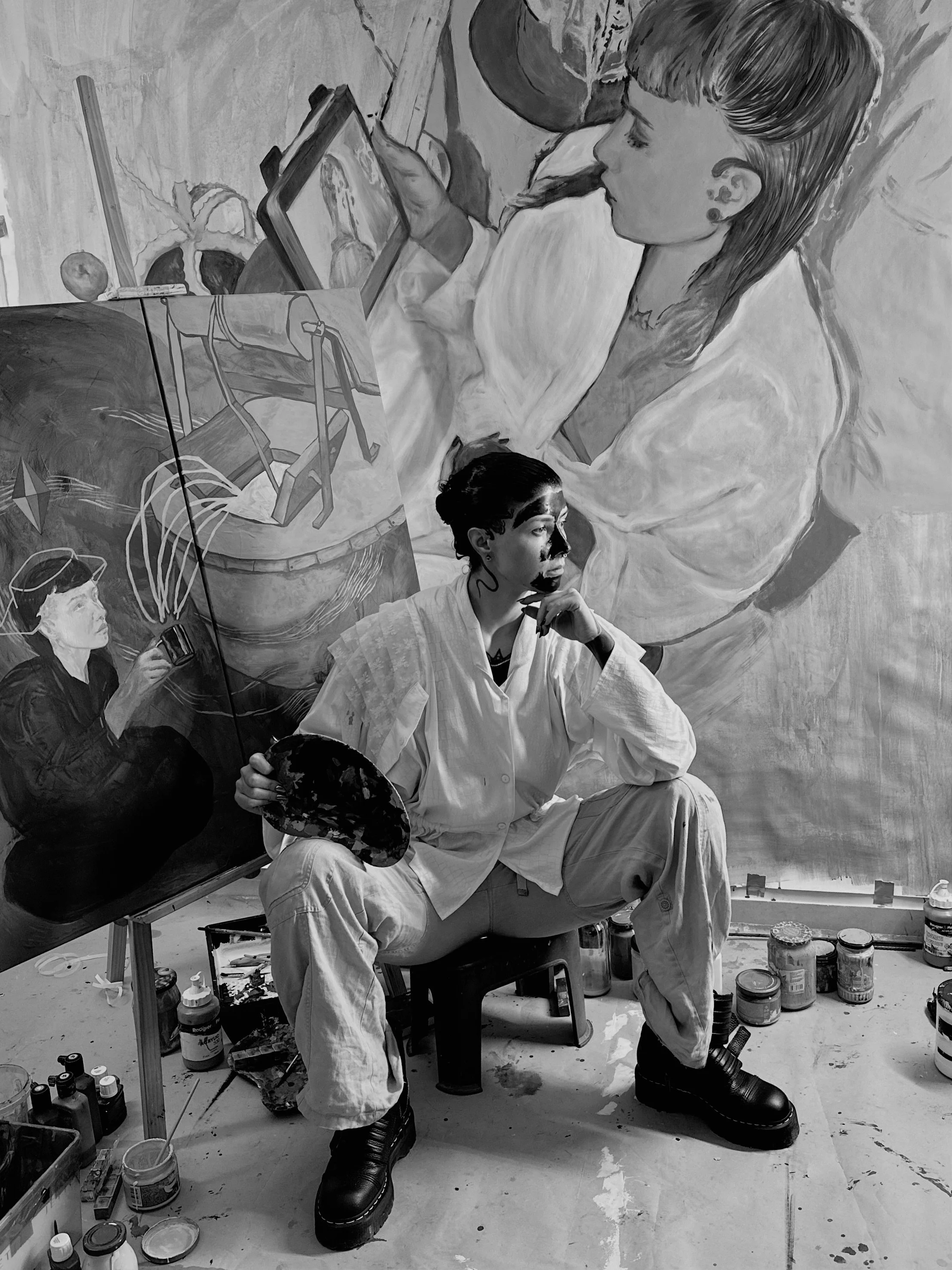 A student artist sitting in front of a large landscape painting of a boat on water, holding a palette and observing his artwork. The artist is dressed in a white coat, with dark hair and boots, surrounded by paints and art supplies in an art studio.