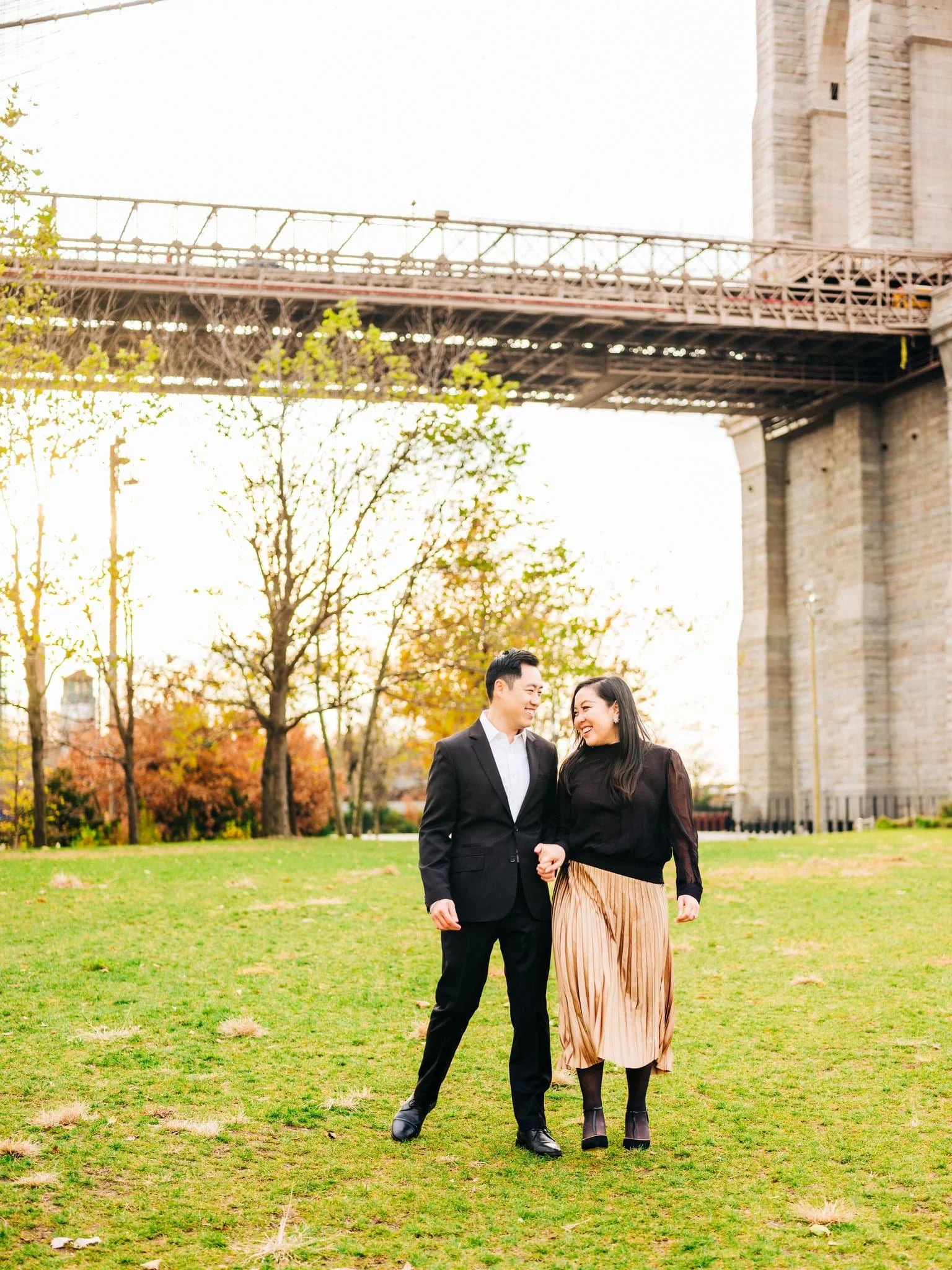 Engaged couple walking through a grassy area with the Brooklyn Bridge behind them during their New York engagement session.