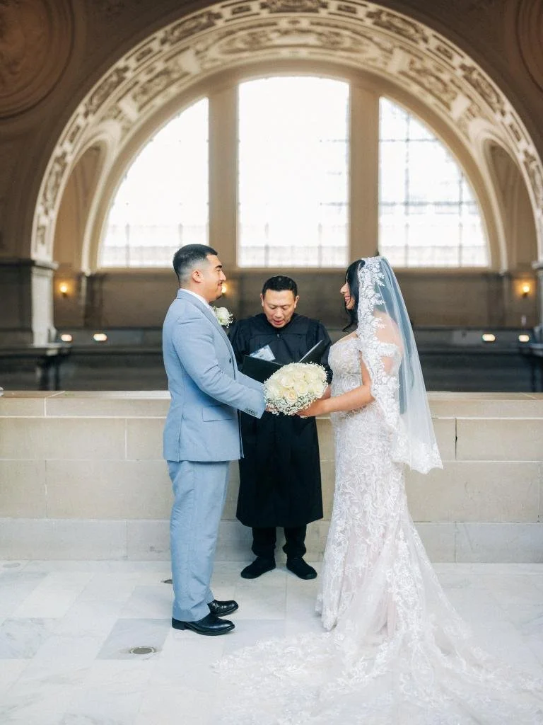 Bride and groom facing each other and holding hands with their officiant during their San Francisco City Hall ceremony