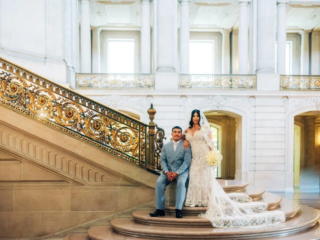 Bride standing beside groom seated at the bottom of the Grand Staircase at San Francisco City Hall