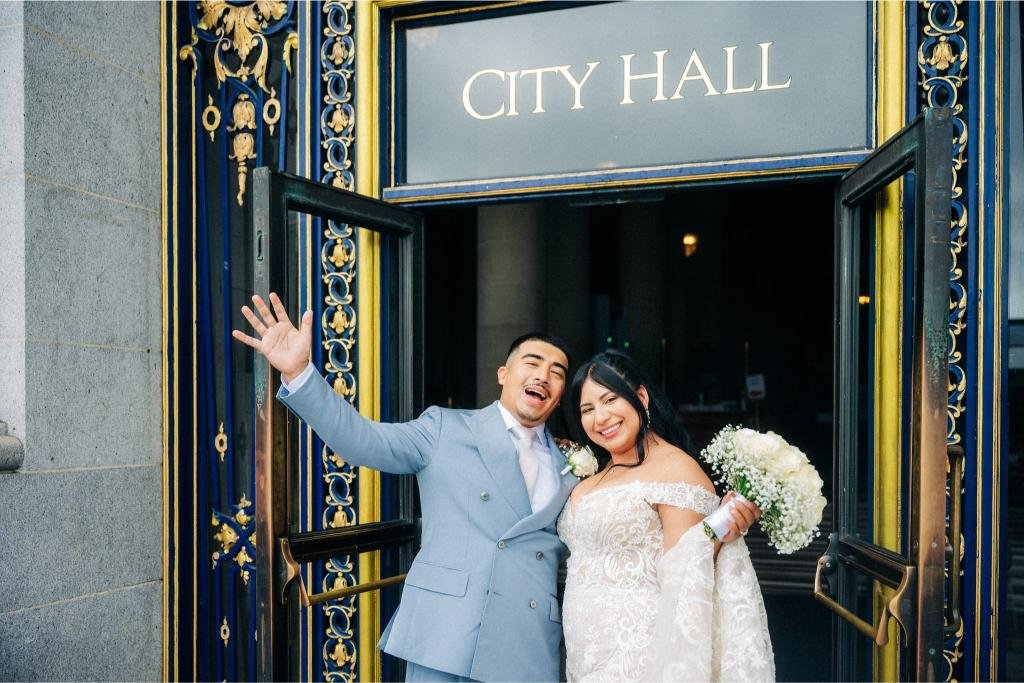 Bride holding her bouquet while the groom raises his hand outside City Hall.