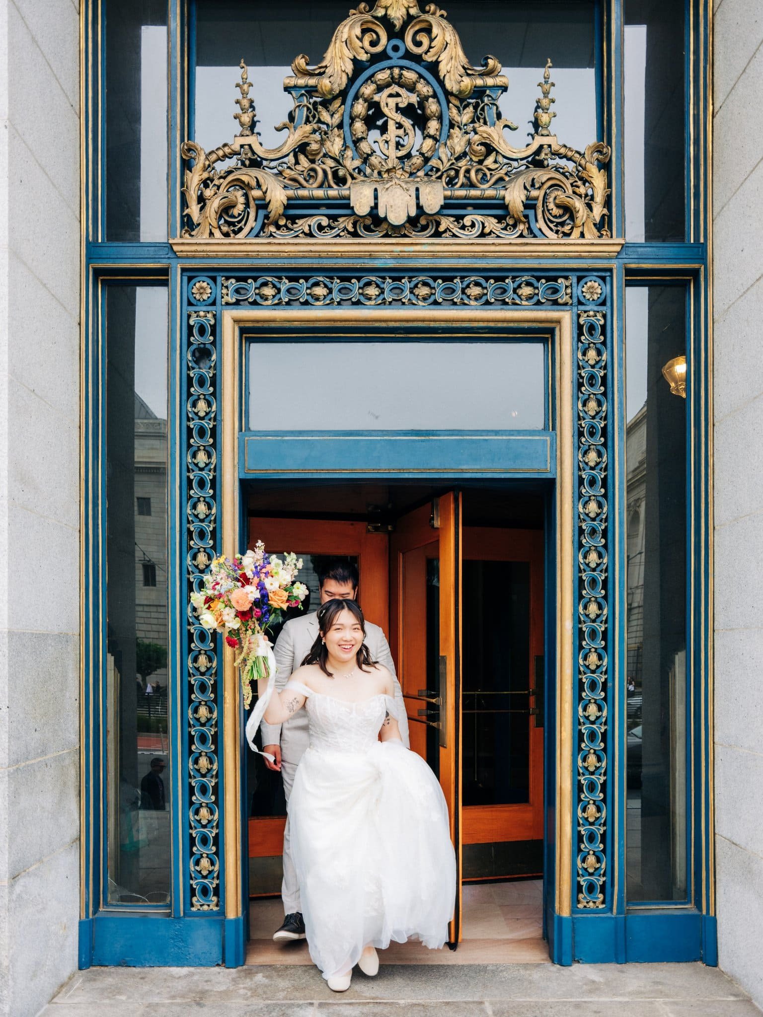 Bride and groom exiting San Francisco City Hall with the bride holding her bouquet above her head in celebration.