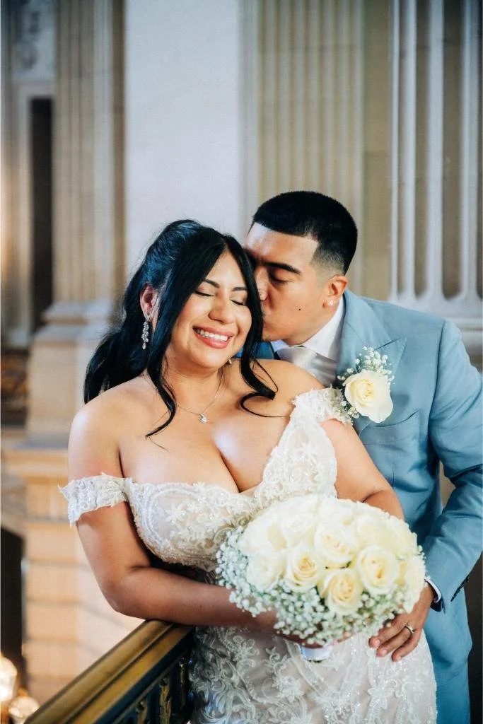 Groom kissing bride on the neck from behind as she smiles inside San Francisco City Hall