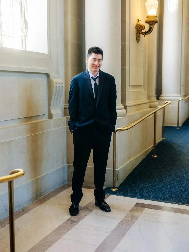 Groom smiling beside a City Hall window with his hands in his suit pockets