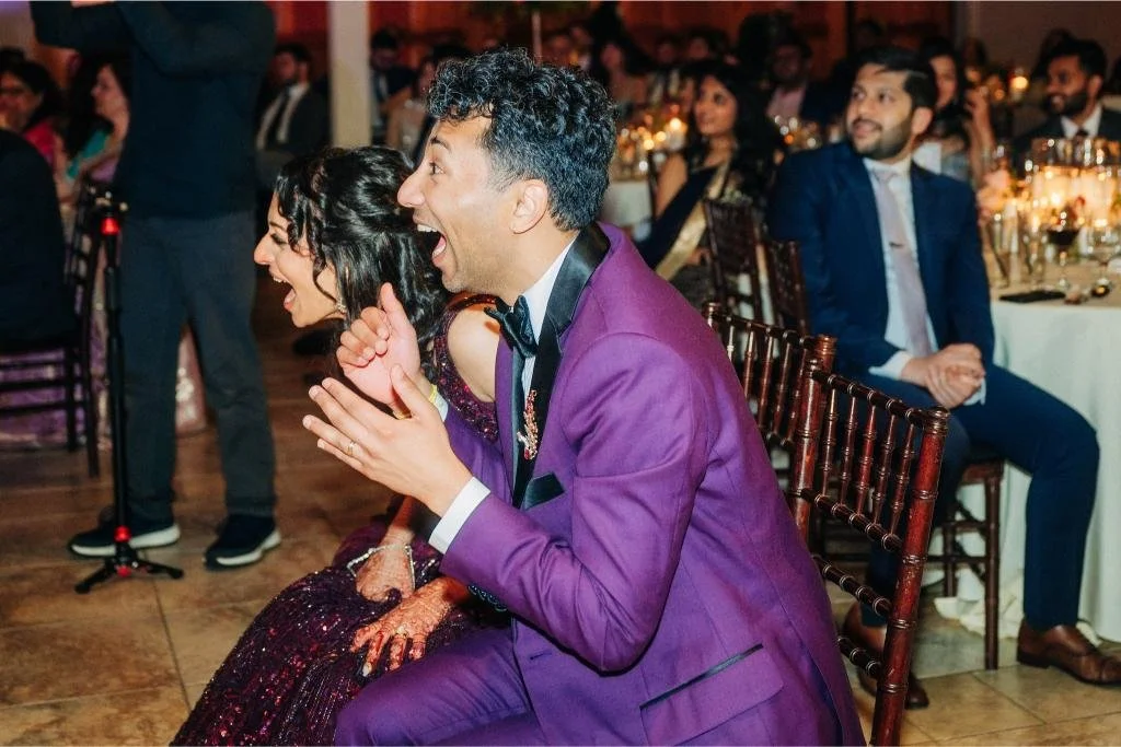 Bride and groom laughing while watching a dance performance by wedding guests