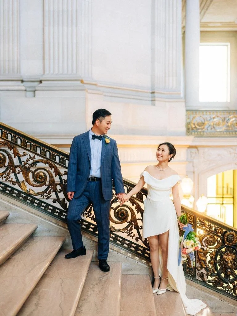 Bride and groom standing on the grand staircase at San Francisco City Hall as he holds her hand and looks at her.