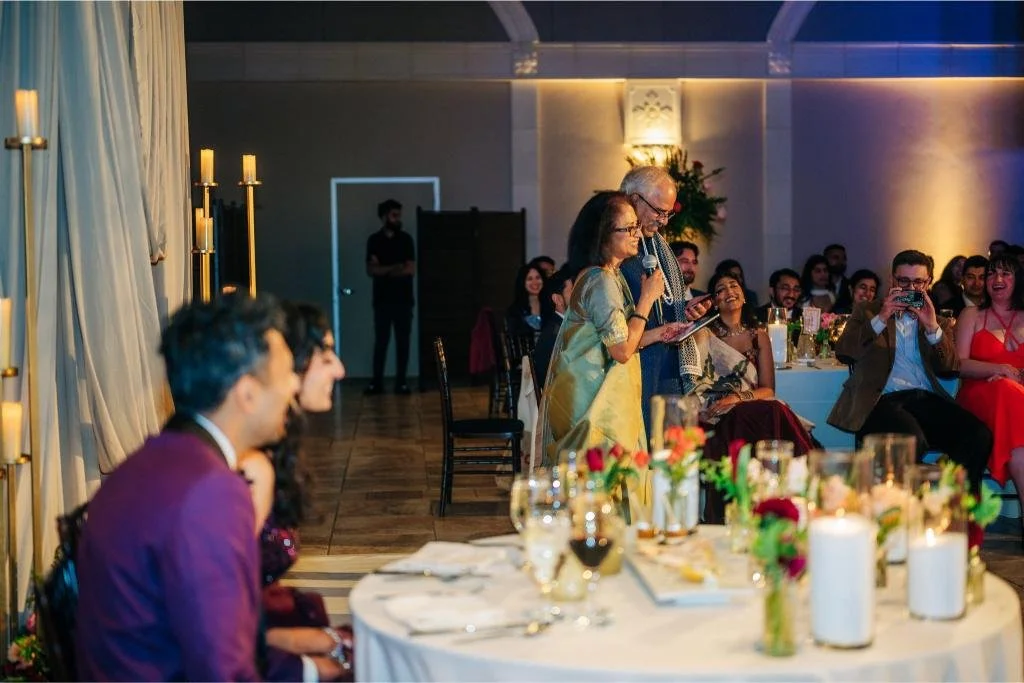 Bride and groom smiling as a wedding guest gives a speech