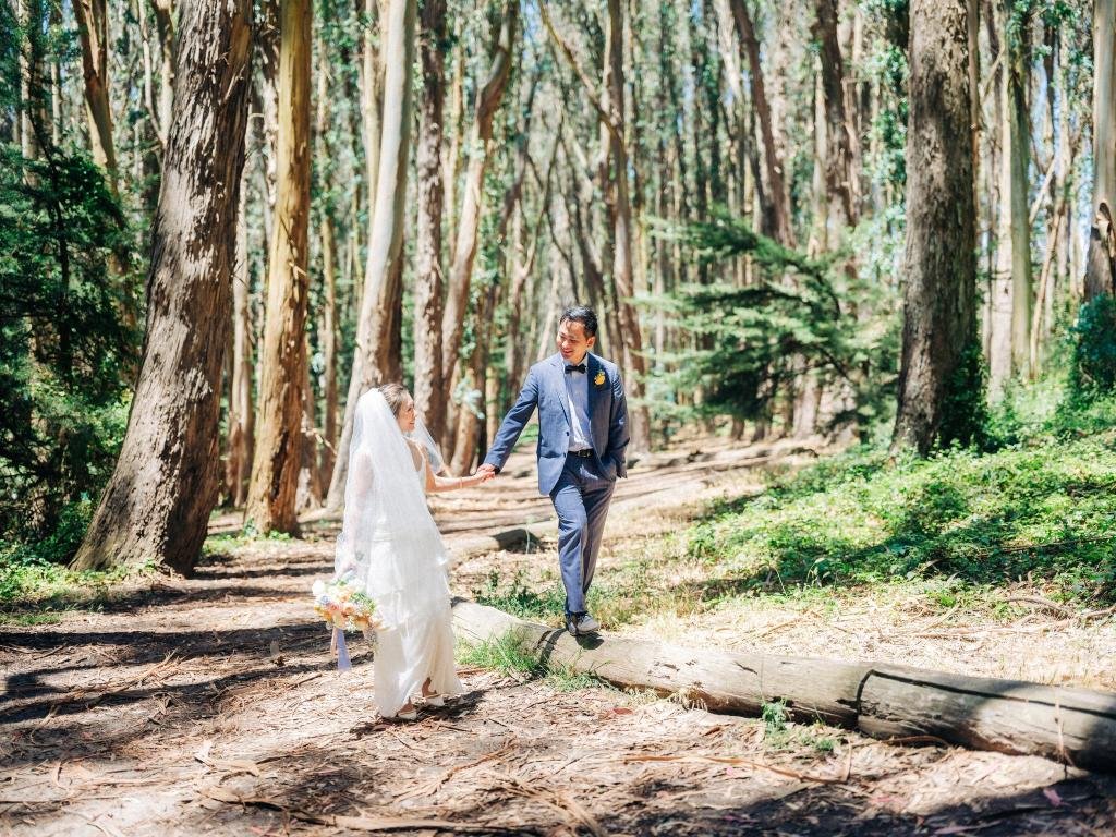 Groom walking along a log while holding the bride's hand as she walks beside him at Lover's Lane in the Presidio.