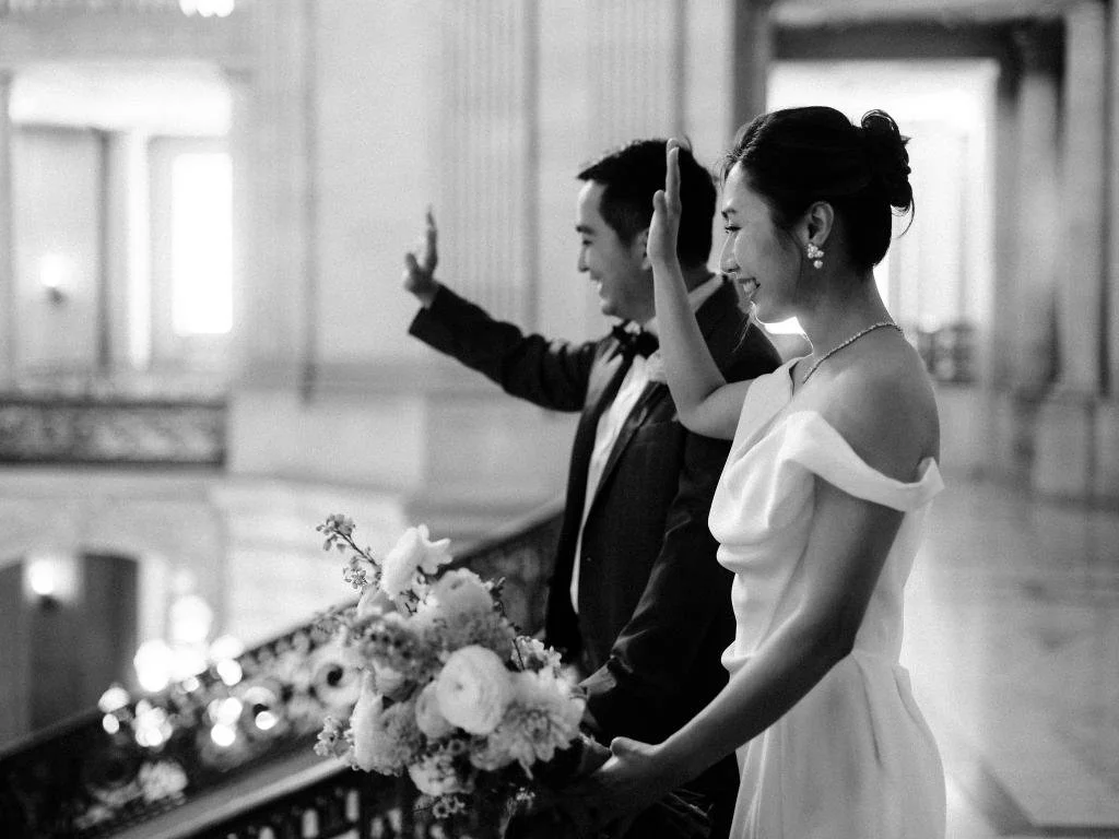 Bride and groom standing above the rotunda at San Francisco City Hall as the bride waves down to their friends.