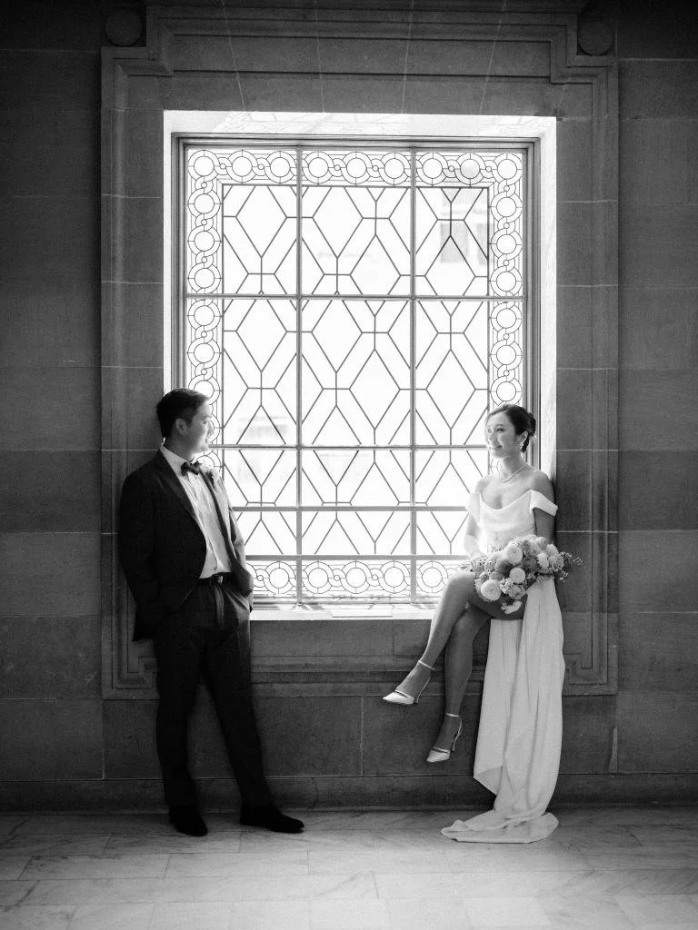 Bride and groom sitting in a large San Francisco City Hall window and looking at each other.
