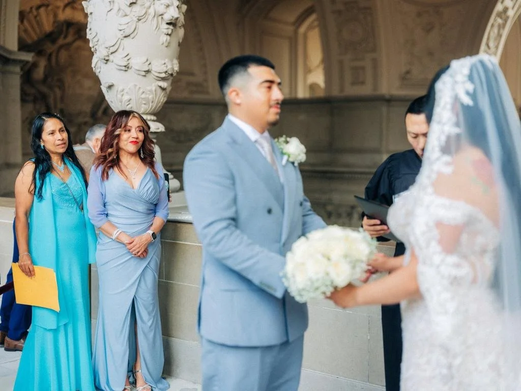 Groom holding bride’s hand while the officiant speaks and guests watch.
