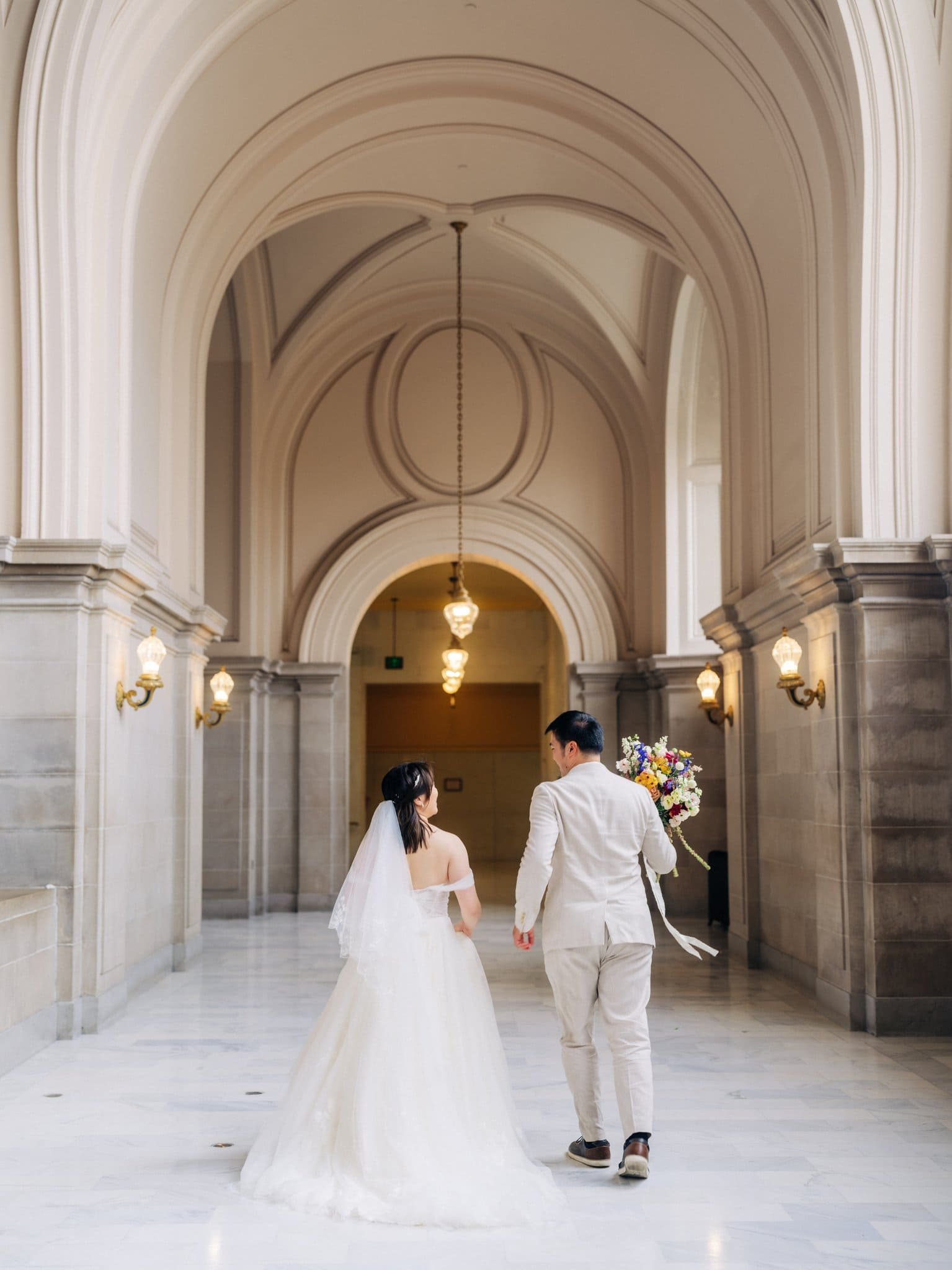 Bride and groom walking hand in hand down a hallway after their San Francisco City Hall elopement ceremony.