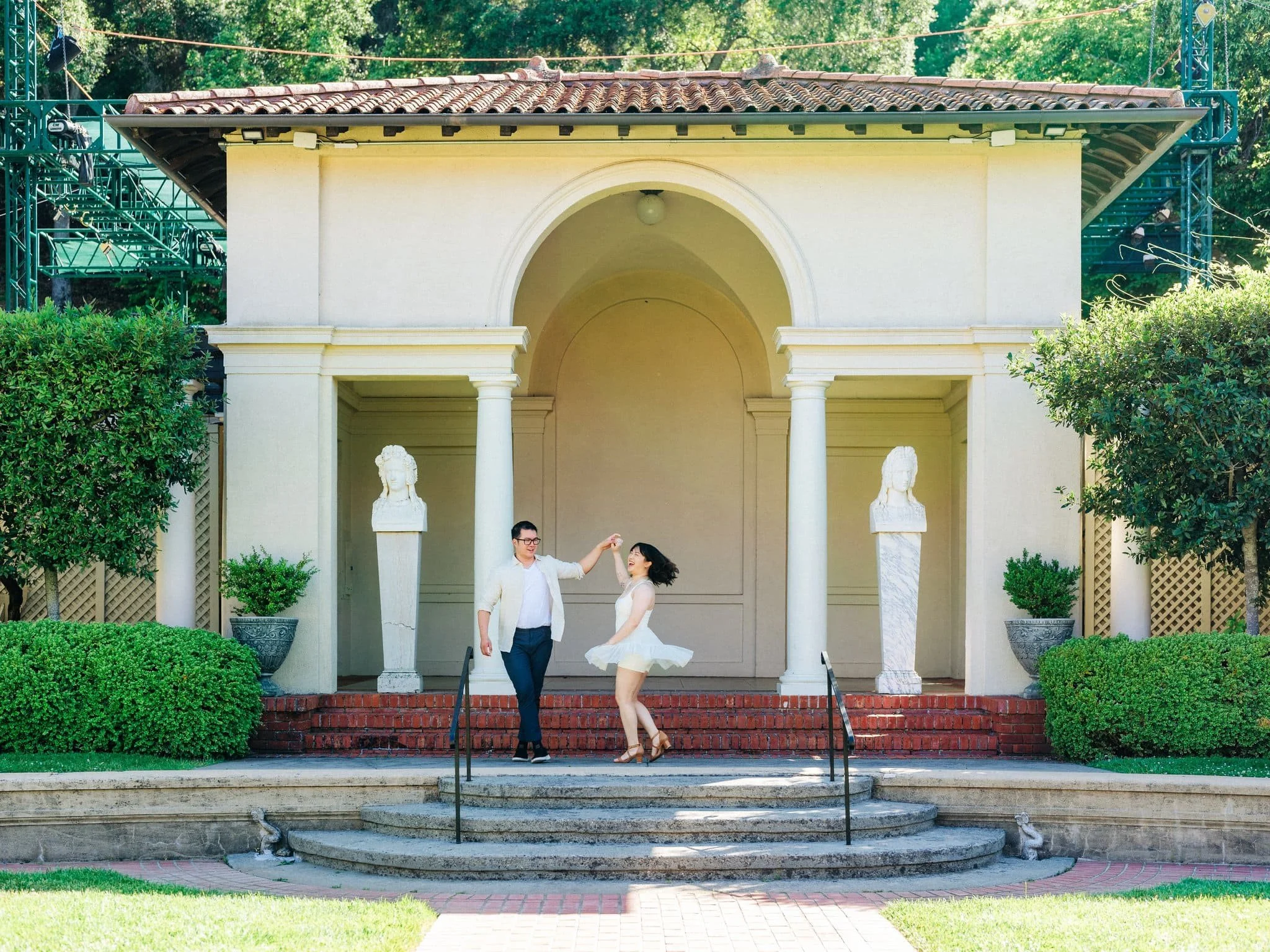 Groom spinning bride-to-be on the staircase at Montalvo Arts Center in Saratoga, with statues on either side.