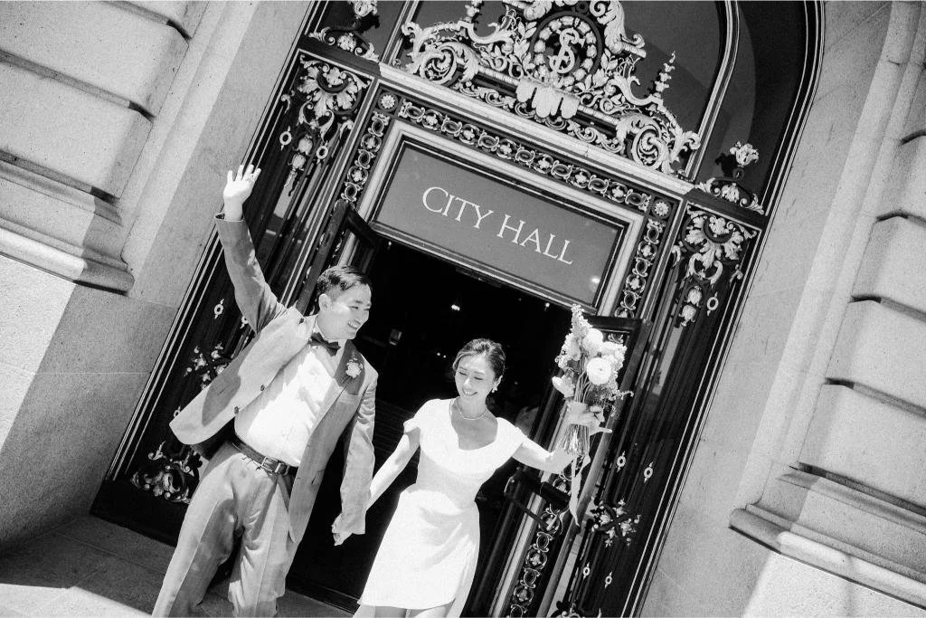 Newlyweds raising their arms in celebration as they leave through the front doors of San Francisco City Hall.