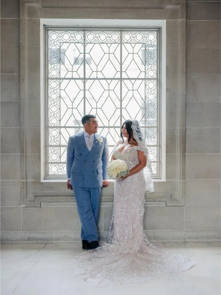 Bride and groom leaning against a window sill beneath a stained glass window at San Francisco City Hall