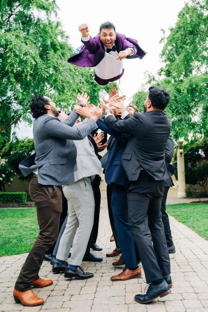 Groomsmen tossing the groom into the air as he strikes a playful Superman pose