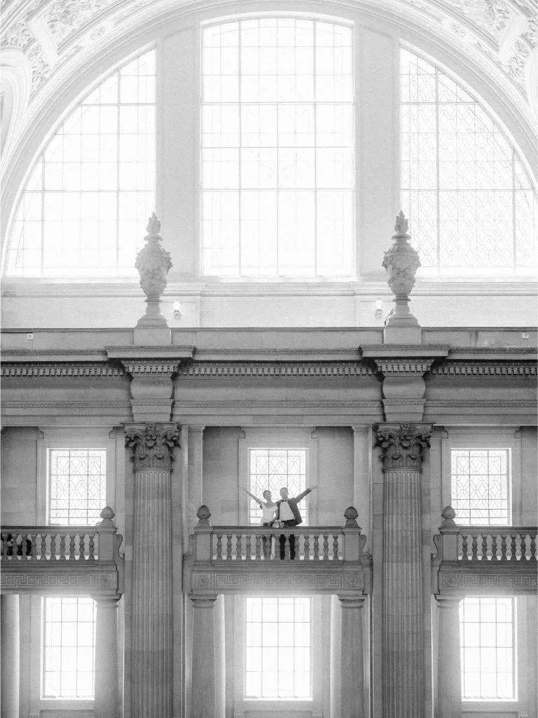 Wide view of San Francisco City Hall with large arched windows and the newlyweds celebrating with their arms raised.