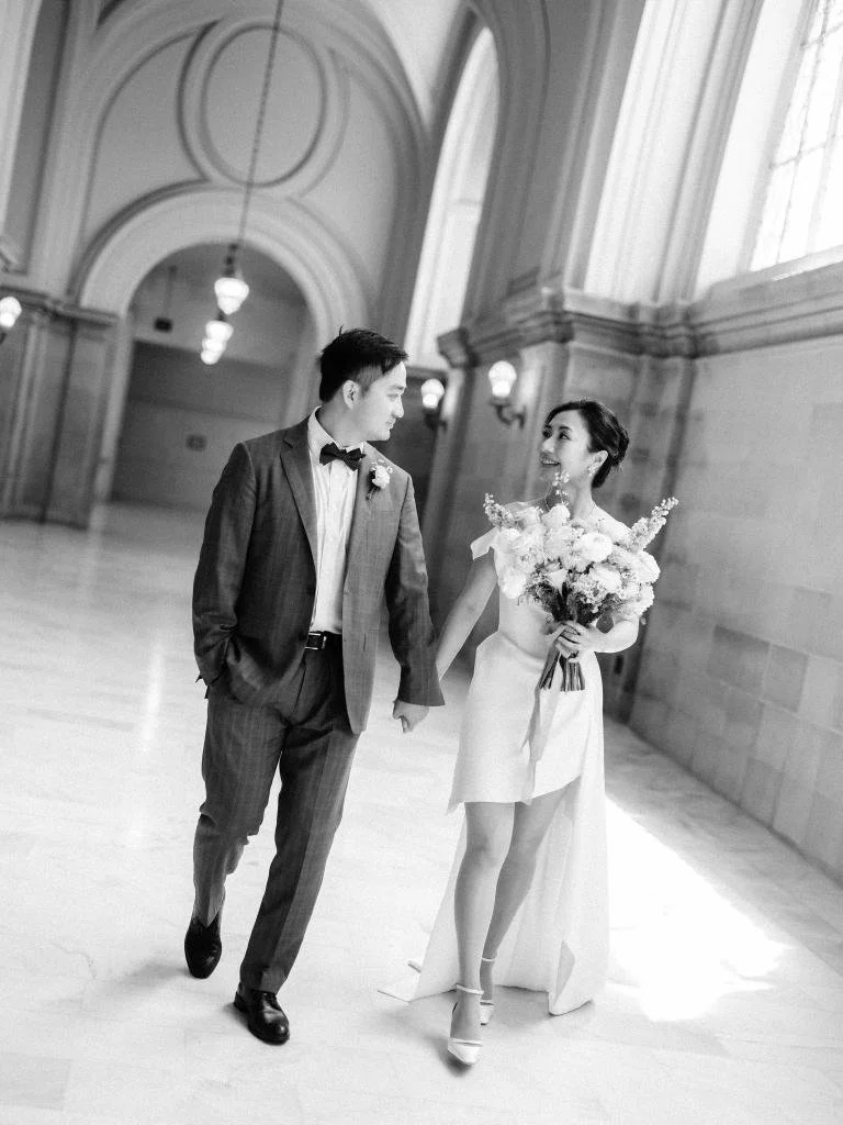 Newlyweds walking hand in hand through San Francisco City Hall while looking at each other.