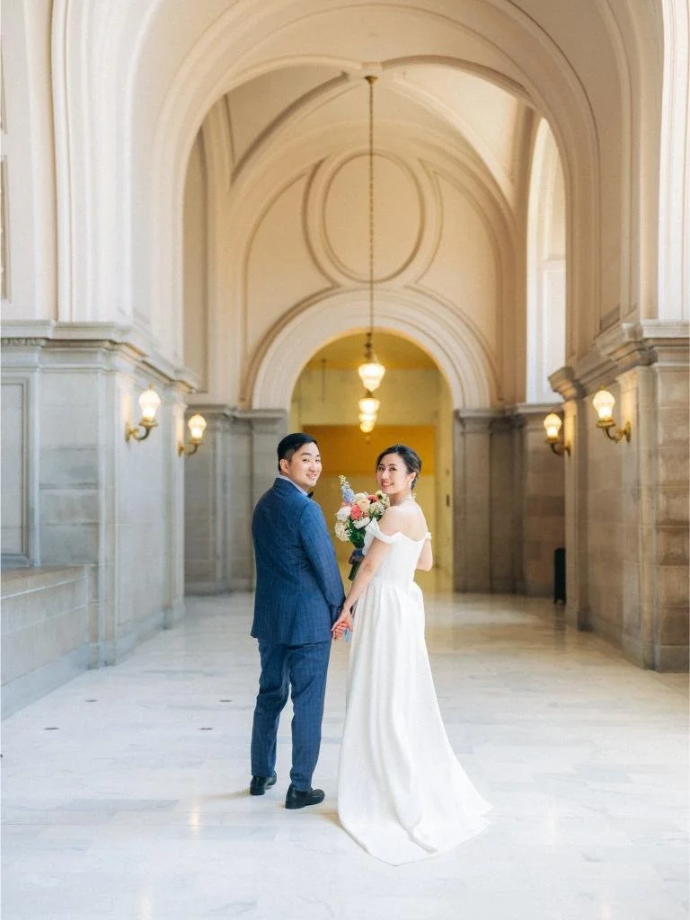 Bride and groom looking back over their shoulders while walking through a San Francisco City Hall hallway.