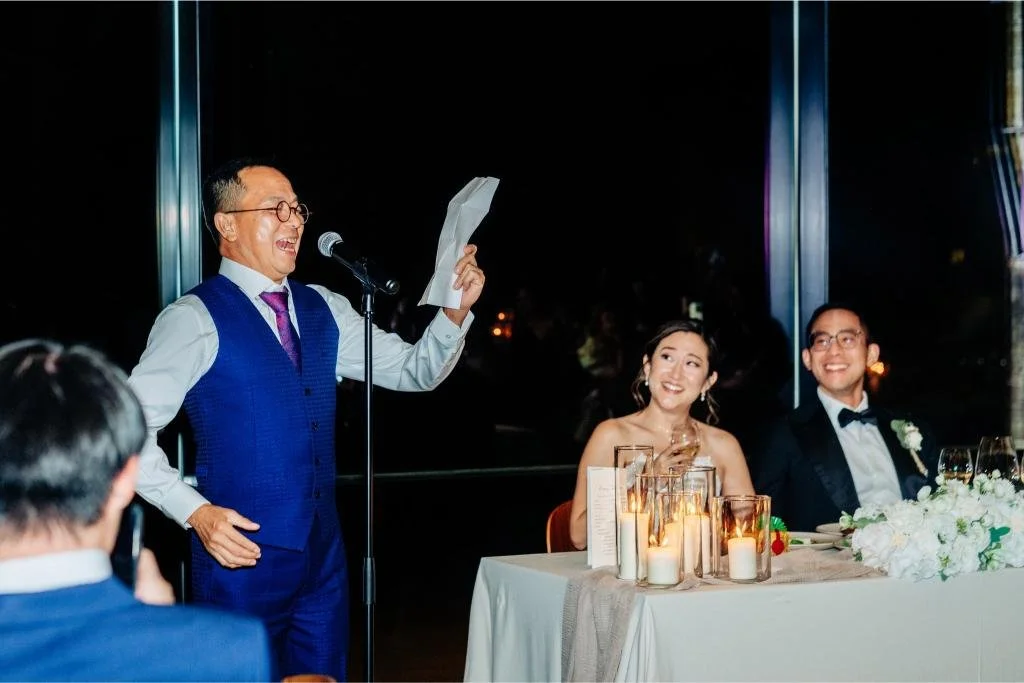 Guest giving a wedding speech as the bride and groom laugh in the background