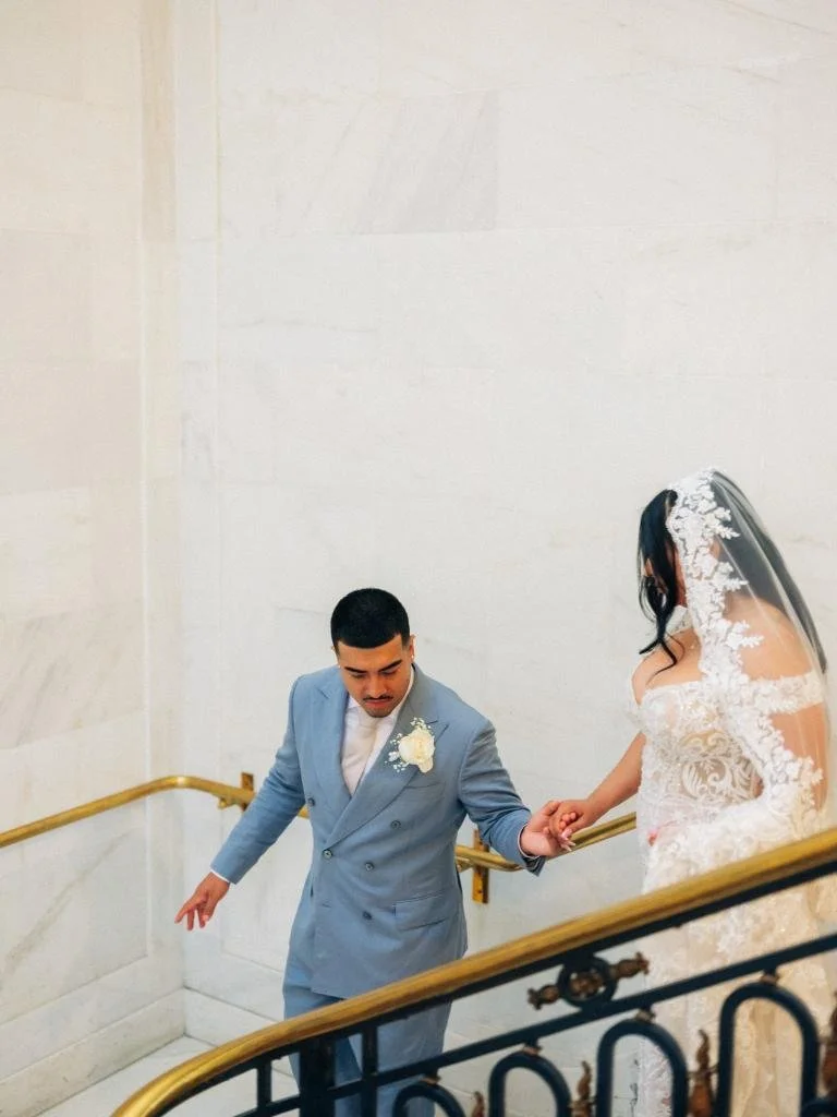 Groom helping bride down the staircase beside the gold handrail.