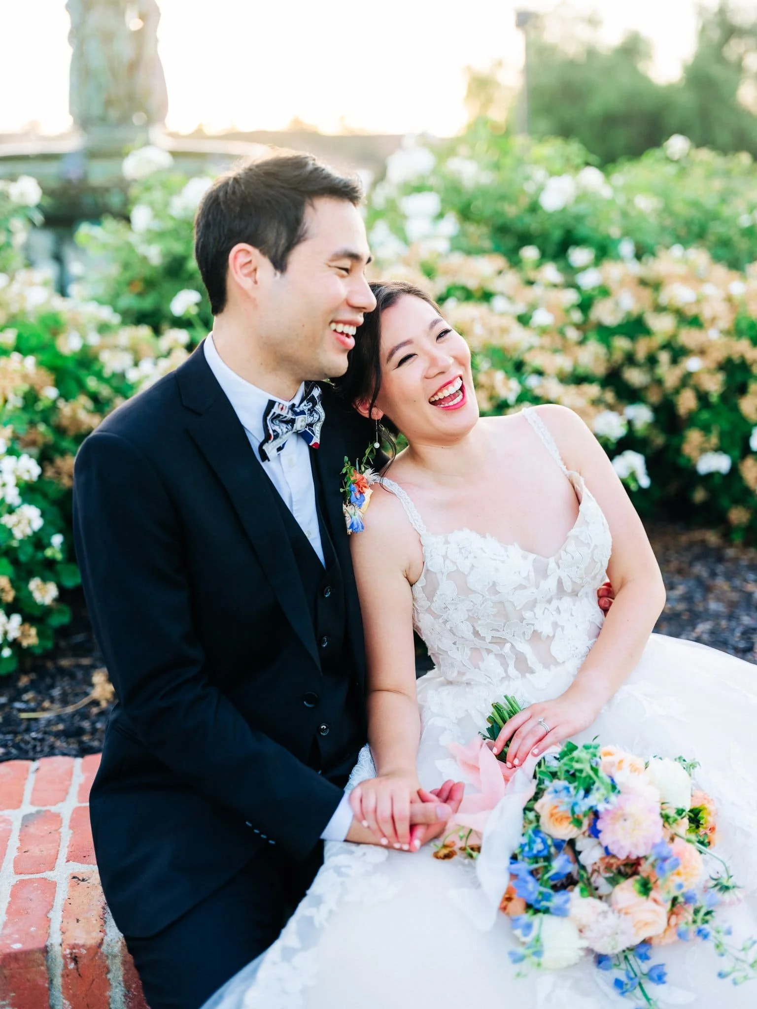 bride and groom sitting and laughing together with flowers behind them palm event center pleasanton wedding
