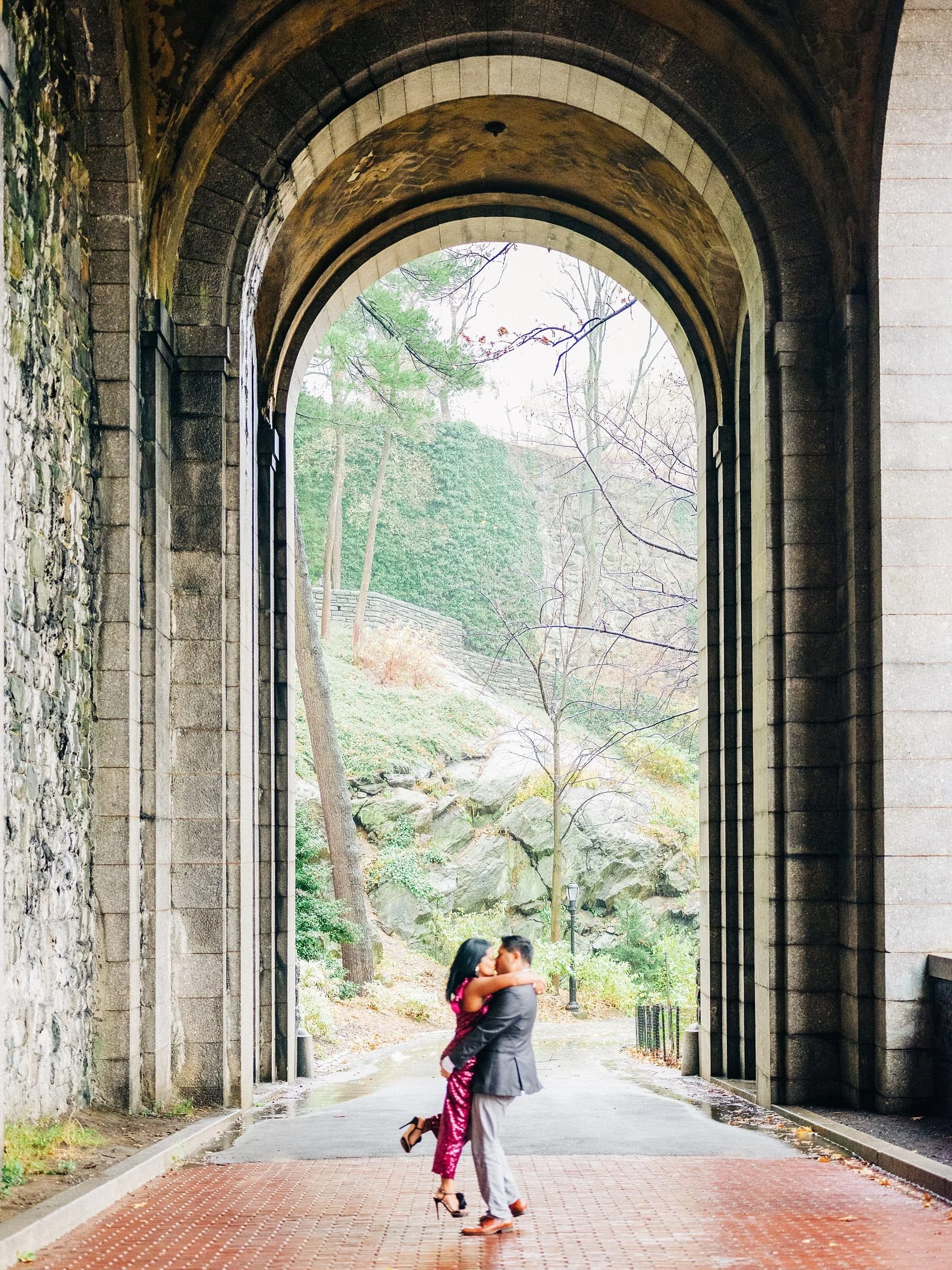Groom lifting bride-to-be under stone arch at Fort Tryon Park during NYC engagement session