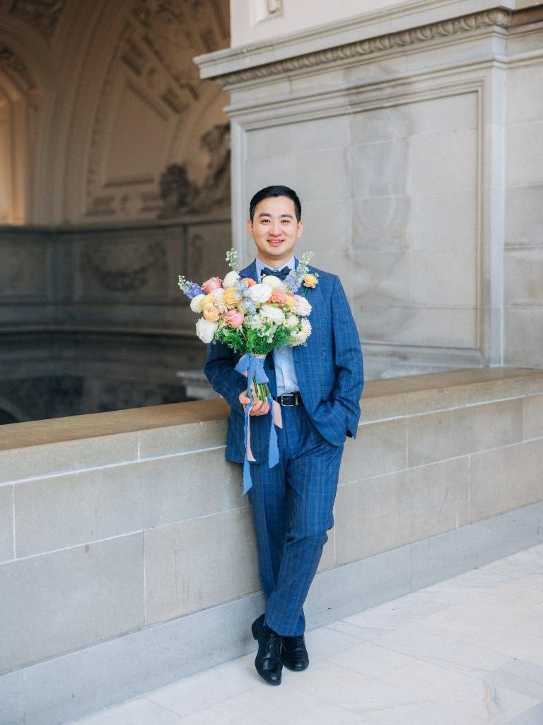 Groom in a blue suit posing with the bridal bouquet during portraits at San Francisco City Hall.