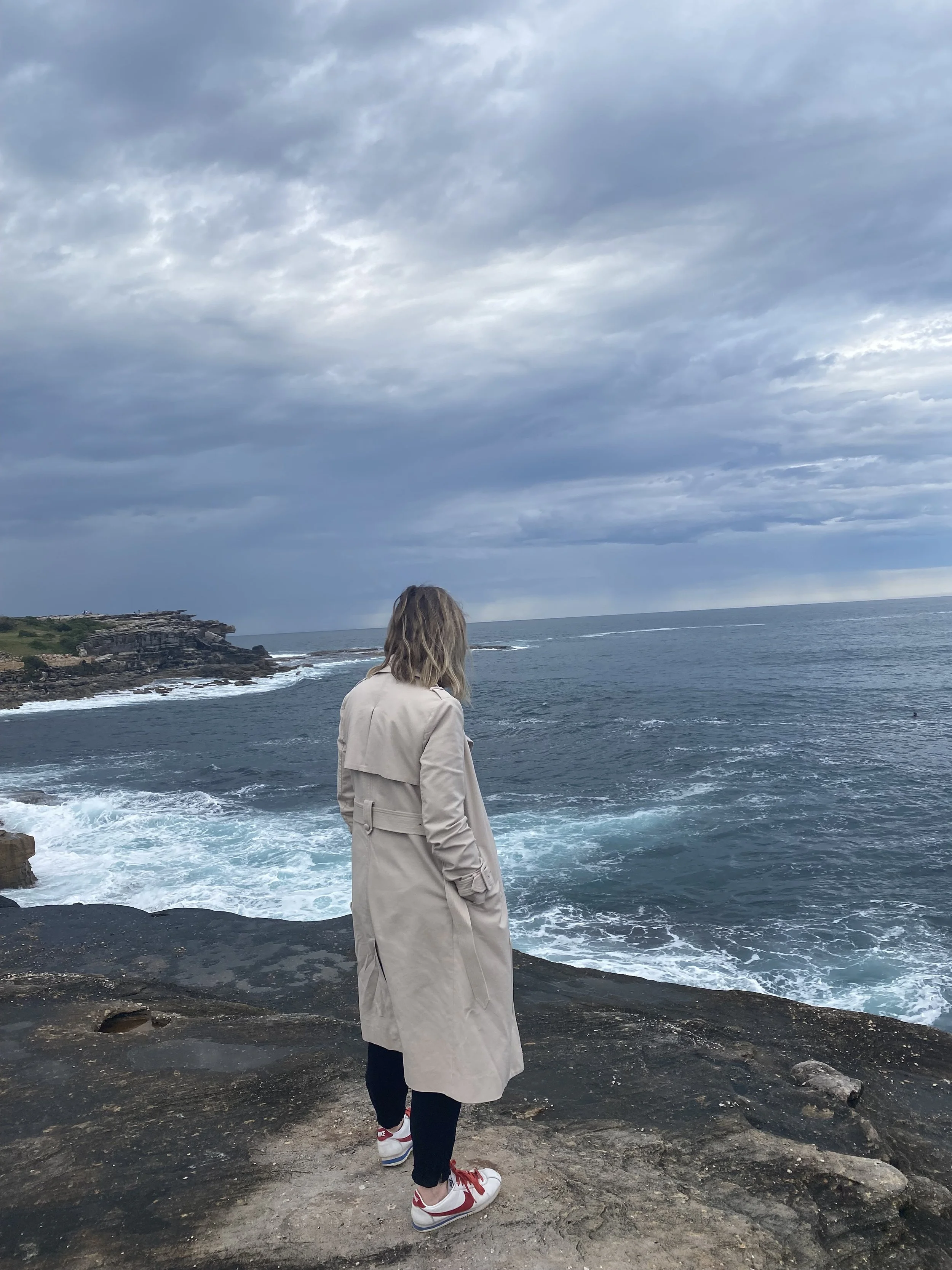 Jet standing on rocky coast by the ocean, wearing a beige trench coat, black pants, and white sneakers with red accents, looking out over the water on a cloudy day.