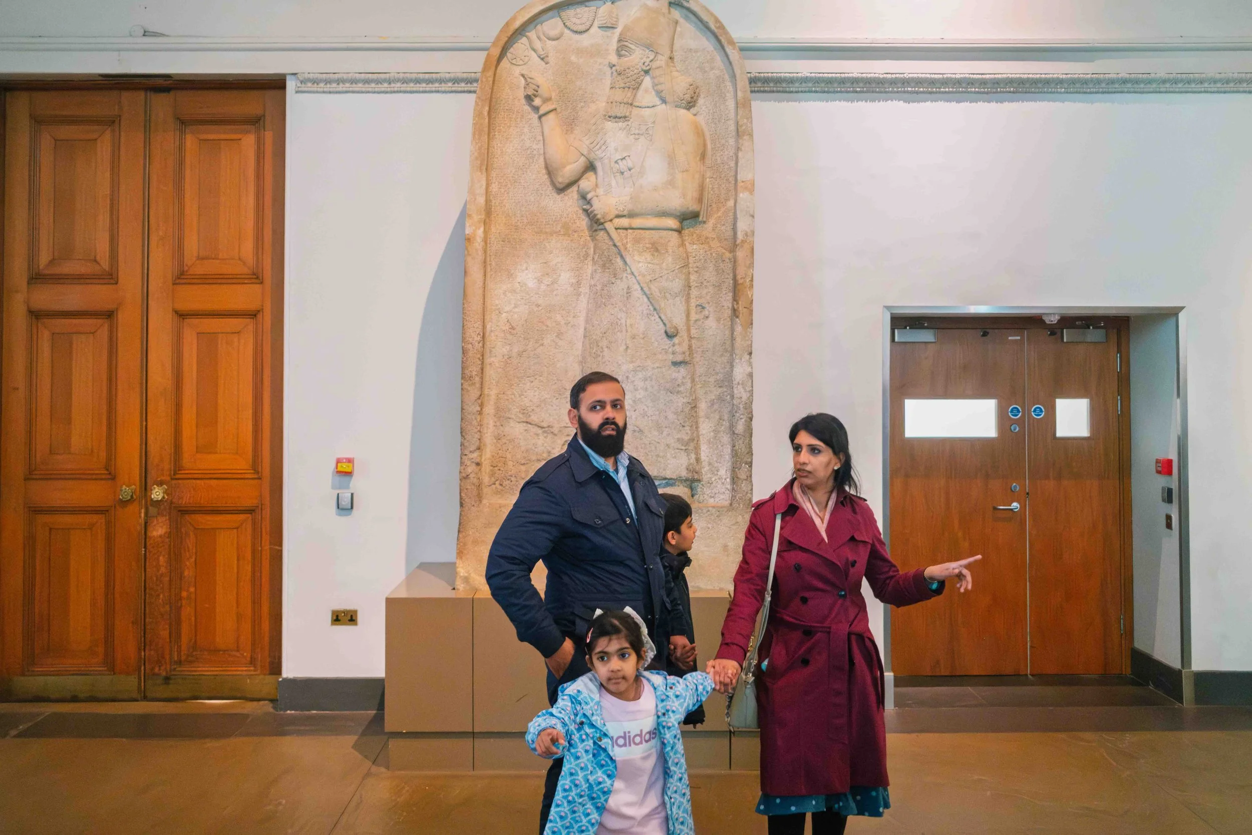 A family of four standing in a museum hallway with a large ancient stone relief sculpture of a man on the wall behind them. The man in the sculpture is holding a staff and appears to be wearing a headdress.