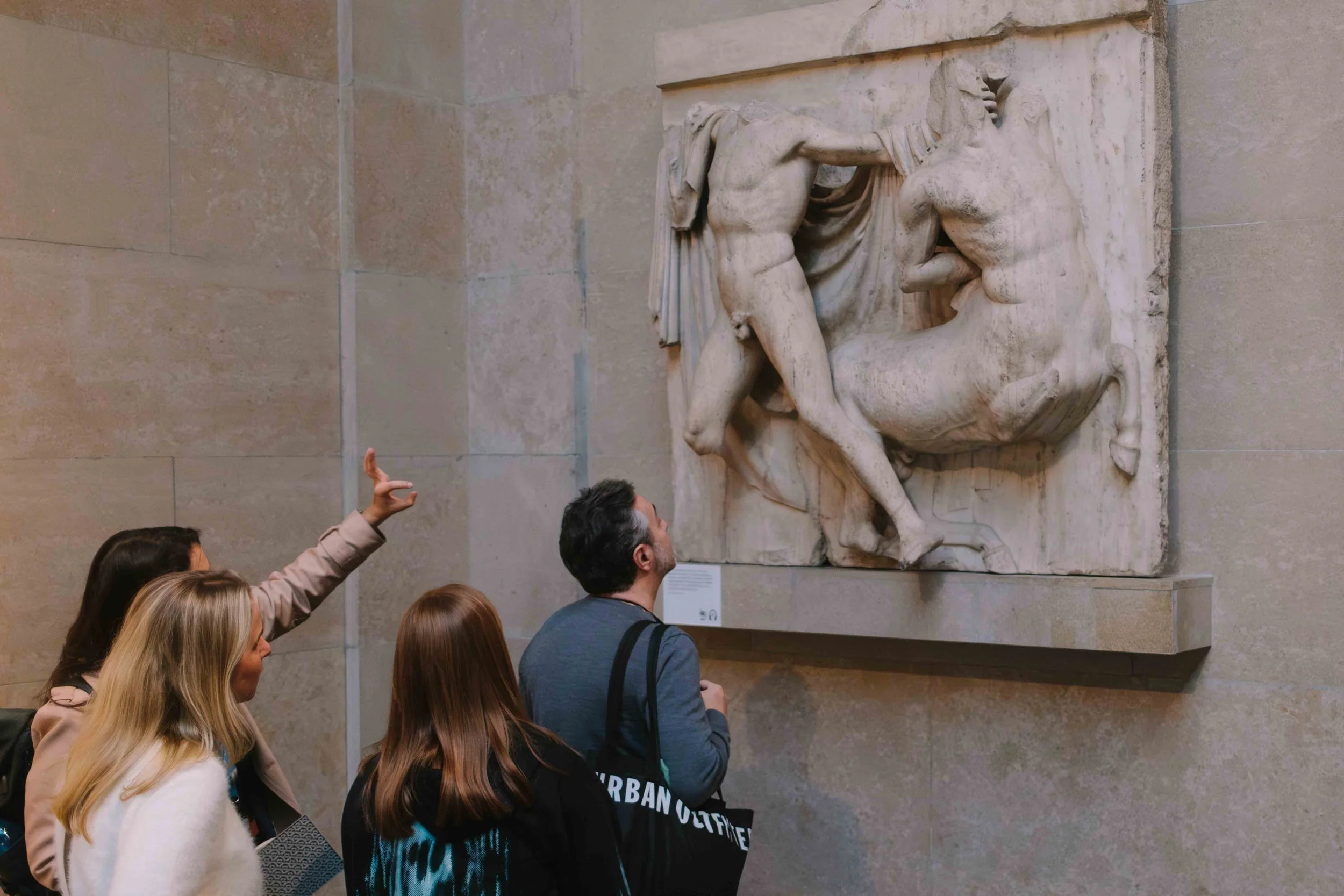 A group of four people observing a marble sculpture of a mythological scene in a museum. One person is explaining while others are listening attentively.