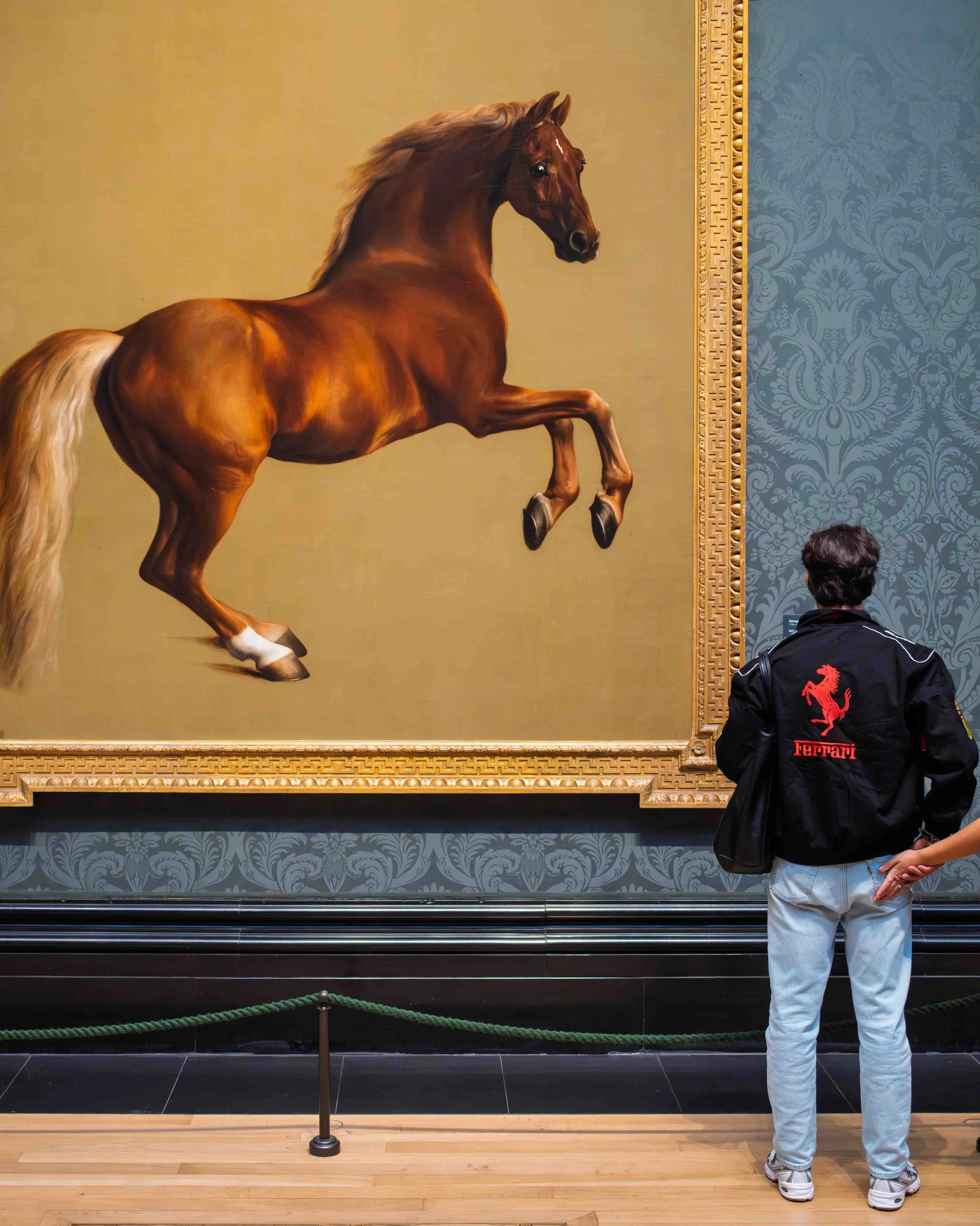 Person wearing a black jacket with a red Ferrari logo, standing in front of a large painting of a rearing horse, inside a gallery or museum with ornate frame and patterned blue wallpaper.