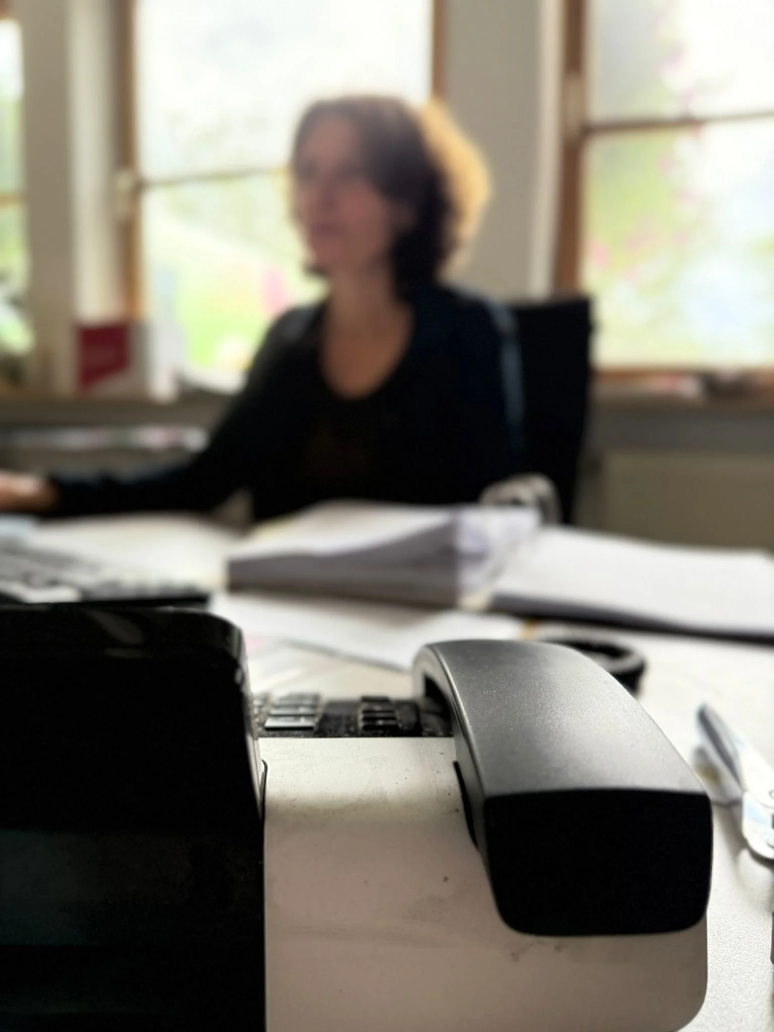 Schreibtisch mit Telefon, Maus, Tastatur und Notizen im Vordergrund, im Hintergrund eine Frauenfigur bei der Arbeit, unscharf.