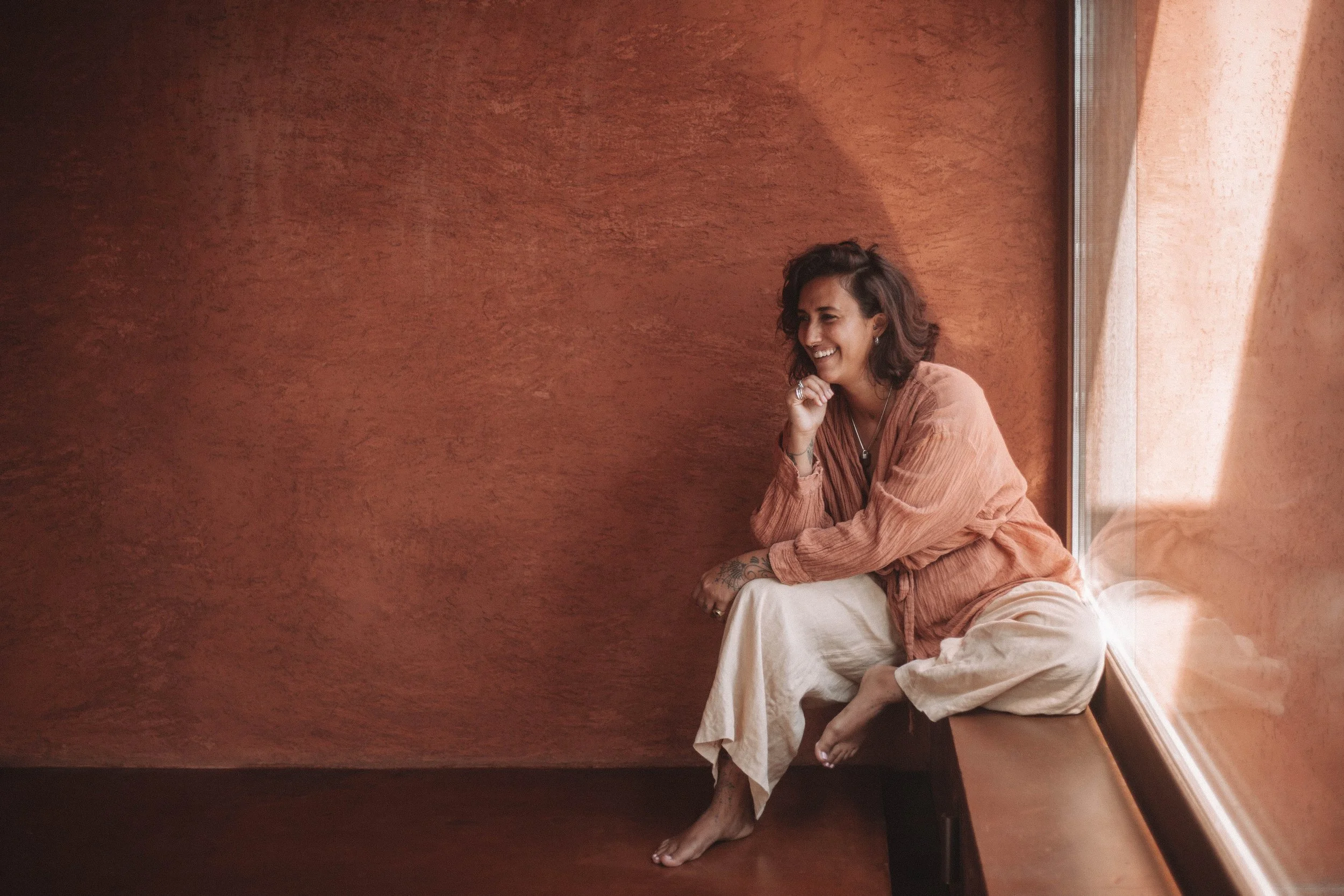 A woman with dark hair, wearing a light brown and beige outfit, sitting on a windowsill by a window with sunlight shining in, smiling and looking to her right.