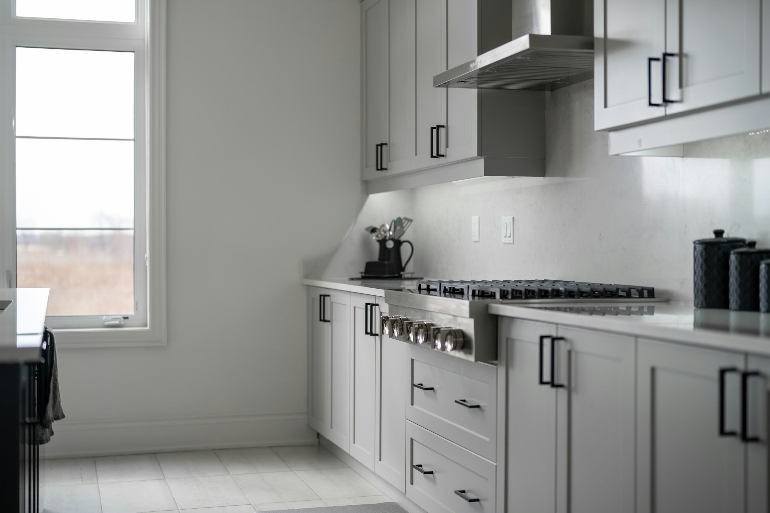 Modern kitchen with white cabinets, a stainless-steel gas stove, and black handles. There are black canisters and a knife block on the countertop, and a window providing natural light.