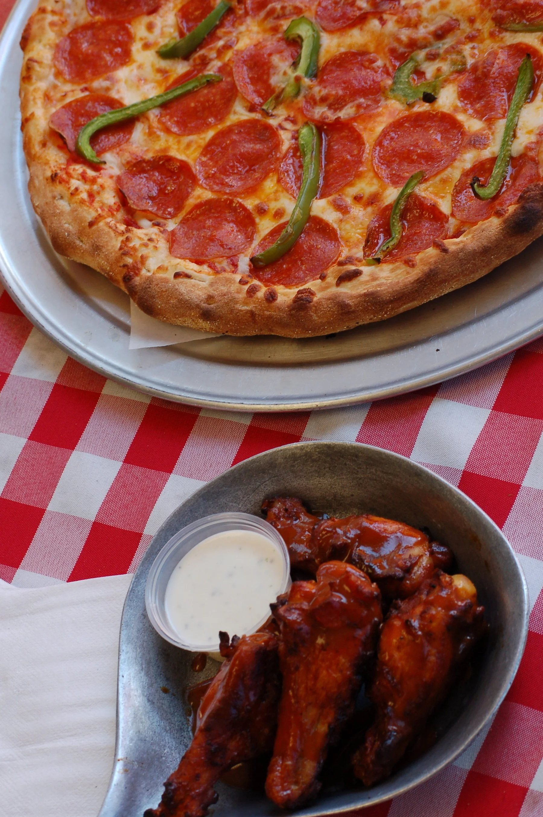 Pepperoni and green pepper pizza on a silver tray, with buffalo chicken wings and ranch dressing in a white bowl, on a red and white checkered tablecloth.