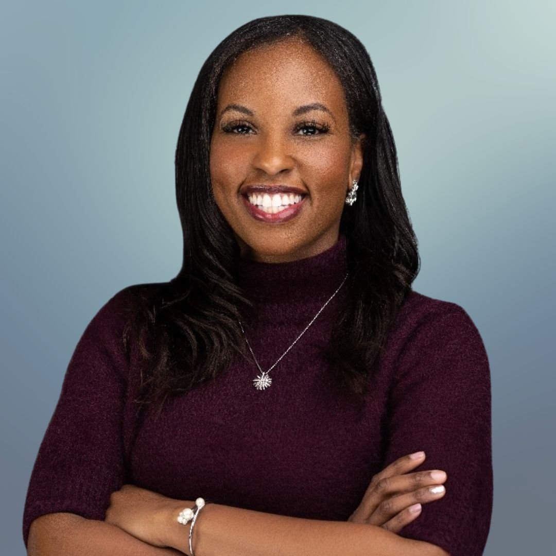 Smiling person with folded arms wearing a dark sweater and jewelry on a blurred background.