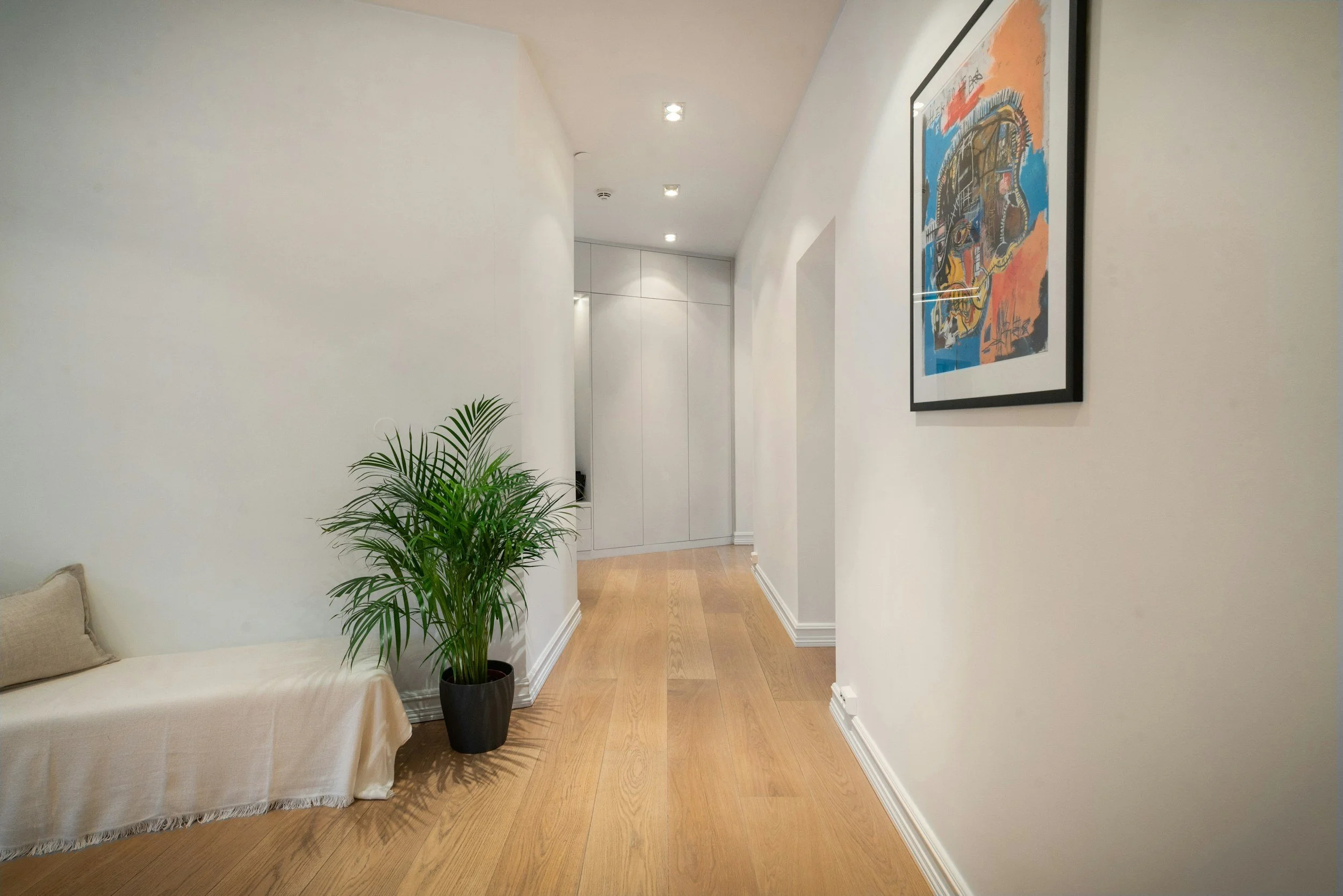 Interior hallway with white walls, a framed colorful abstract painting on the right, a potted green plant in the center, a beige sofa with a pillow on the left, light wooden flooring, recessed ceiling lights, and white baseboards.