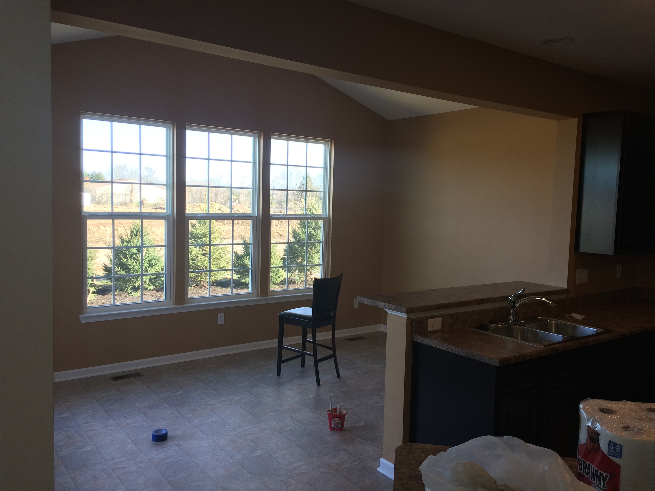 Empty kitchen with three large windows, a chair, a tiled floor, a granite countertop, and some household items on the floor and counter.