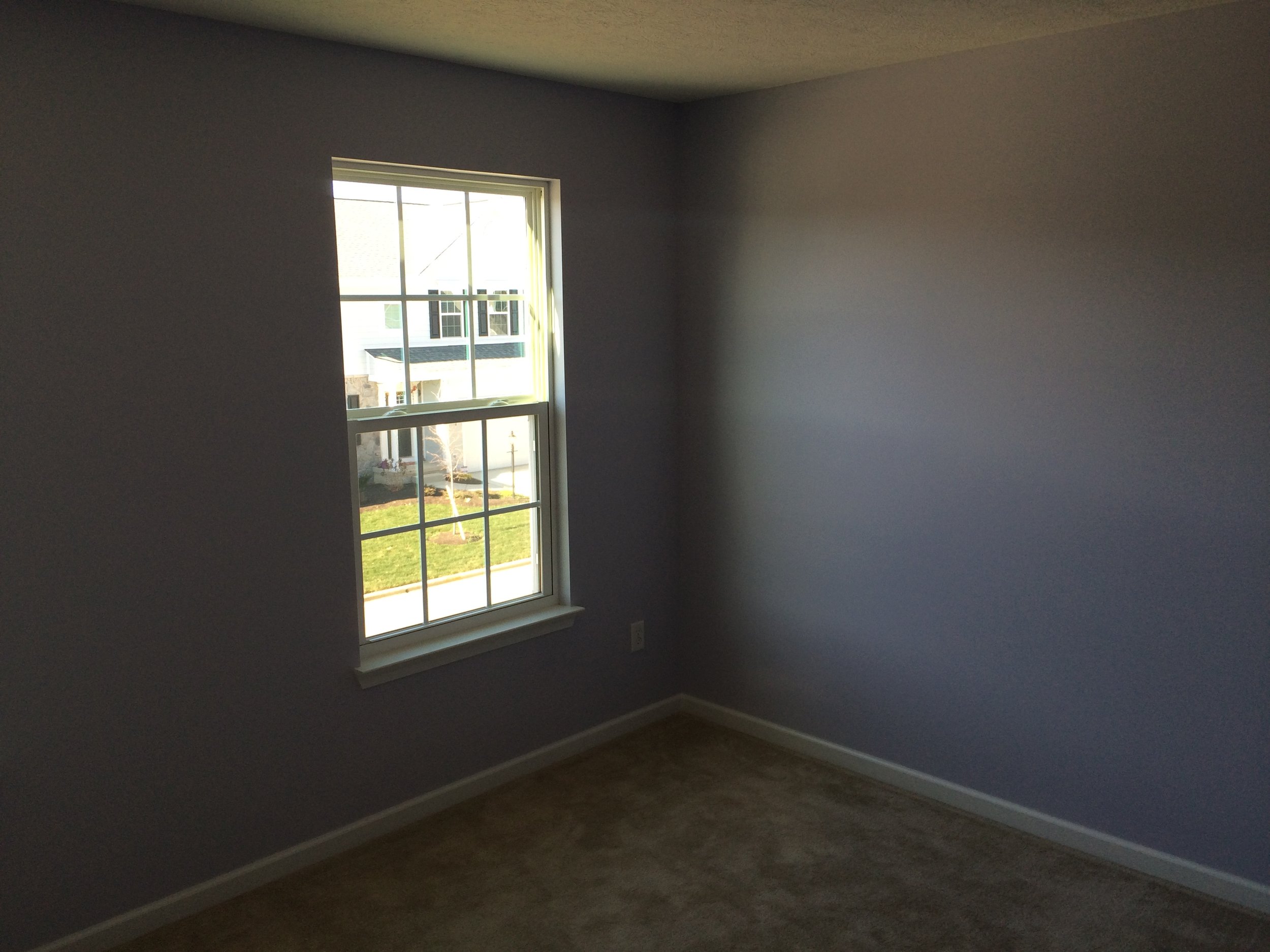 Empty room with gray walls, a window showing a house outside, and a brown carpeted floor.